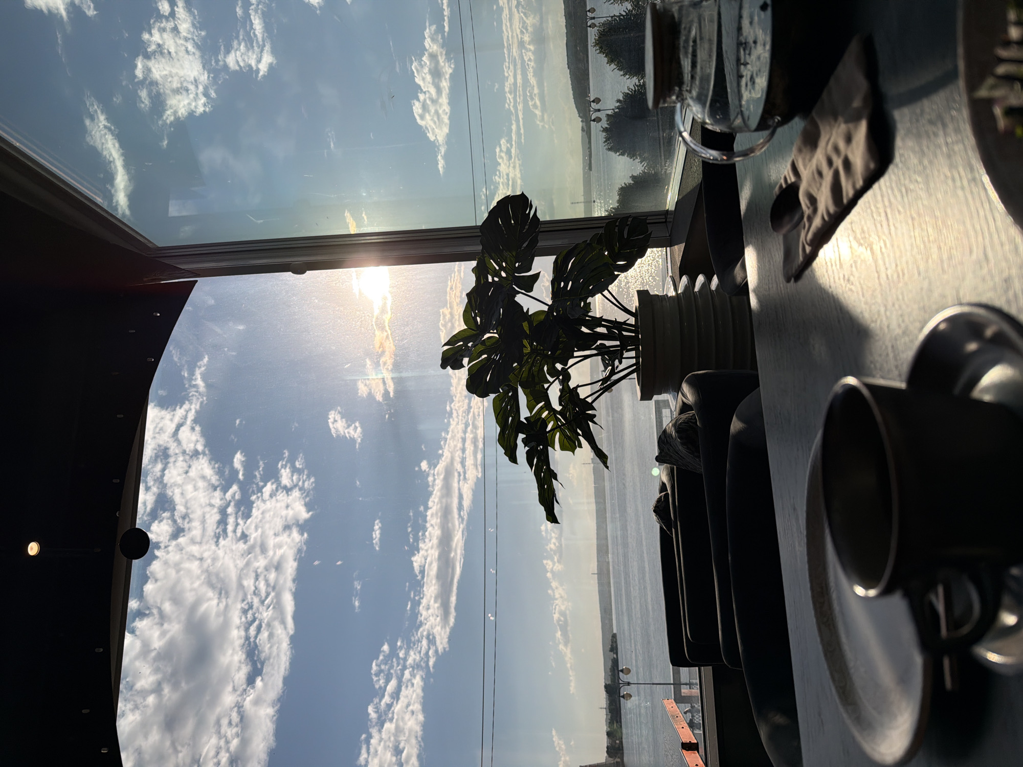 This image depicts a view from a window, likely in a restaurant or cafe, overlooking a body of water. The scene is set during sunset, with the sun casting a warm glow and creating reflections on the water.
In the foreground, a table is set with a dark ceramic mug, a saucer, and a spoon wrapped in a napkin. A glass teapot filled with dark liquid is also visible on the table.
Beyond the table, through a large window, a wide expanse of water is visible, with a distant shoreline. The sky is a mix of blue and white clouds, illuminated by the setting sun.
In the middle ground, there are several dark, plush chairs arranged facing the window, suggesting a seating area. A potted Monstera plant with large, green leaves stands in a white planter, adding a touch of nature to the interior.
The overall impression is one of a tranquil evening setting, likely in a location that offers scenic views. While no specific details in the image definitively place it in Izhevsk, Russia, it is a plausible setting for such an establishment.