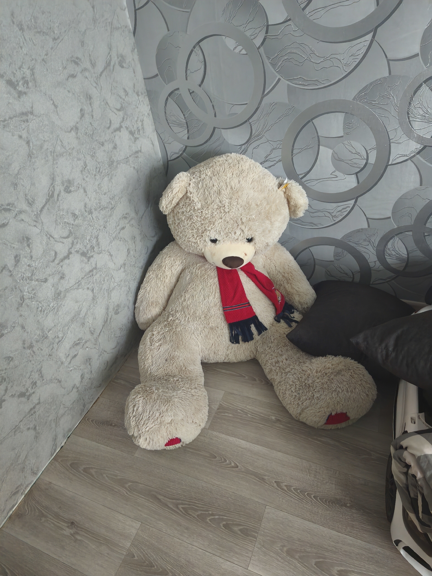 An oversized, light beige teddy bear sits on a light brown wood-grain floor in the corner of a room. The bear is wearing a red and blue scarf around its neck. The wall behind the bear has a textured grey pattern. To the right of the bear, two dark grey pillows are visible, along with a portion of a white object with a folded grey and white checkered blanket on top, possibly a stroller or a bed. The room appears to be indoors, with no external environmental features visible.