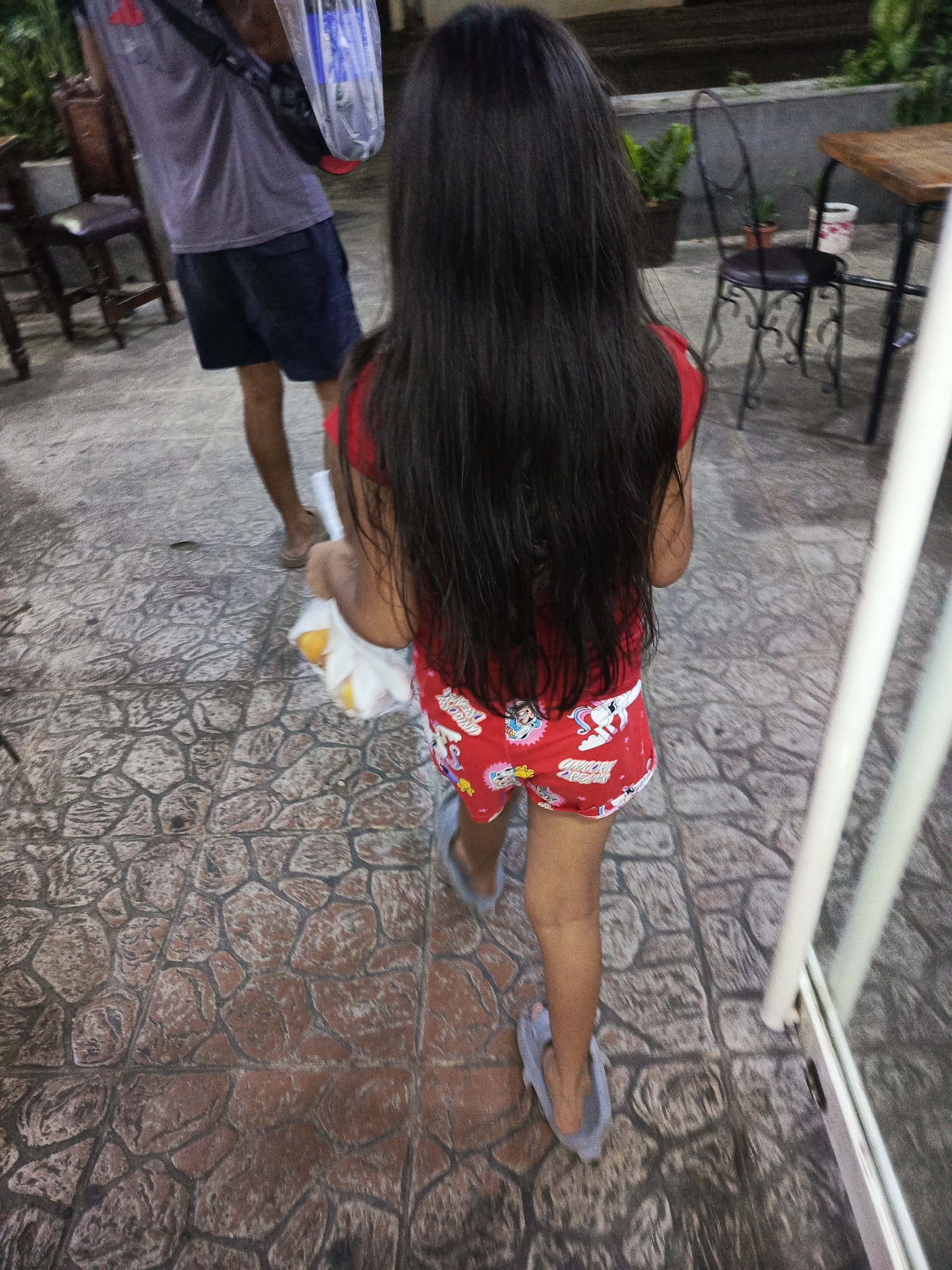 A young girl with long, dark hair is walking on a stone-paved surface alongside an adult male. She wears a red shirt, cartoon-printed shorts, and blue slippers, carrying a white plastic bag in her left hand. The man is wearing a gray t-shirt, dark blue shorts, and carrying a bag of bottled water. The setting appears to be an outdoor area, with chairs and tables visible in the background, along with some potted plants. Based on the reference to Altagracia, Venezuela, it is likely that this scene is taking place in that city.