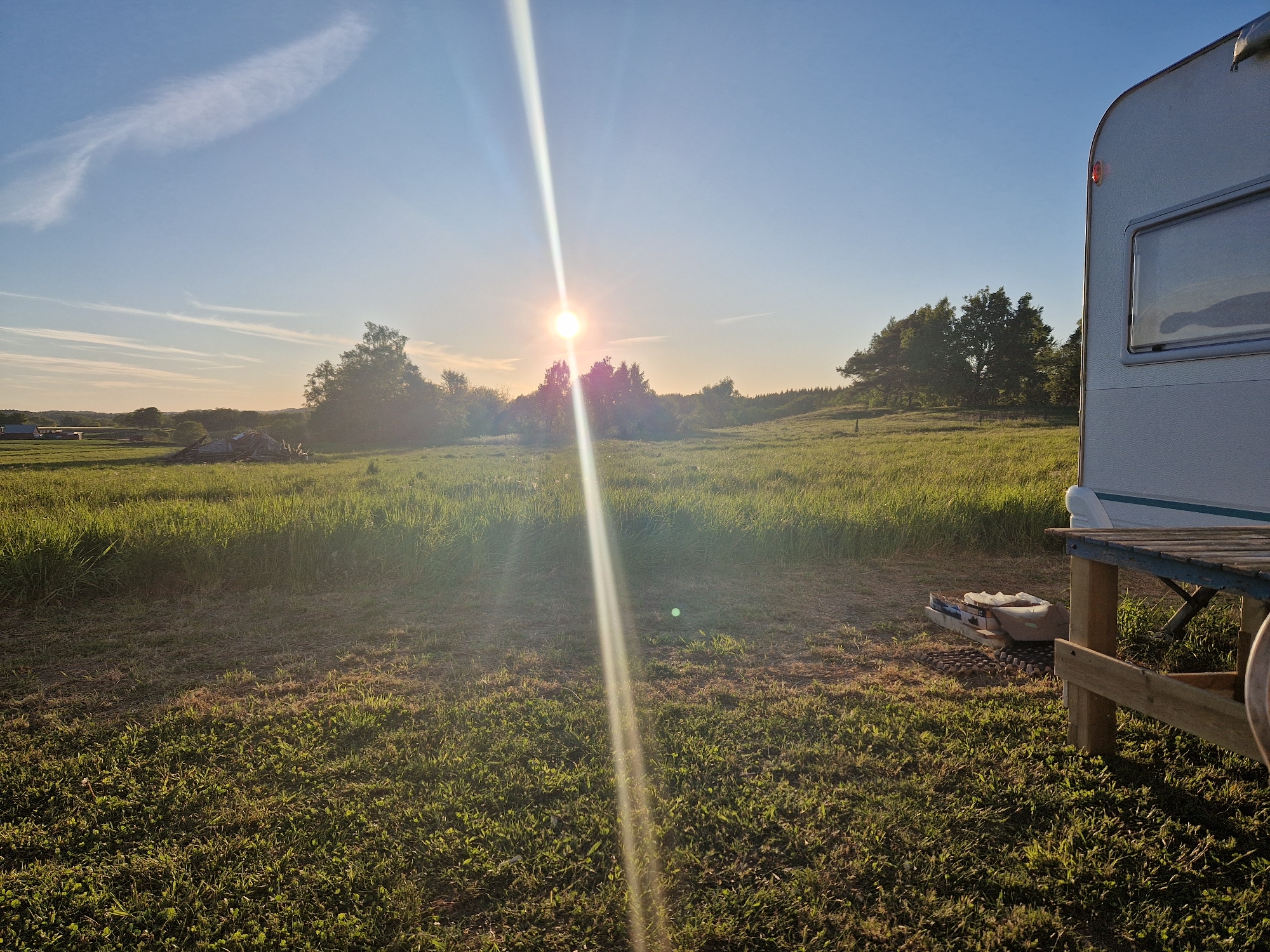 The image depicts an outdoor scene in Vessigebro, Sweden, characterized by a grassy field under a bright sky with the sun prominently shining. The sun is positioned centrally, emitting a strong beam of light downwards. A caravan is visible on the right side of the frame, with a wooden table extending from it. Miscellaneous objects, including what appears to be gardening equipment, are situated beneath the table and beside the caravan. The field has varying heights of grass and vegetation, suggesting a natural or semi-maintained landscape. In the background, trees are visible, delineating the horizon. The overall environment suggests a rural or countryside setting.