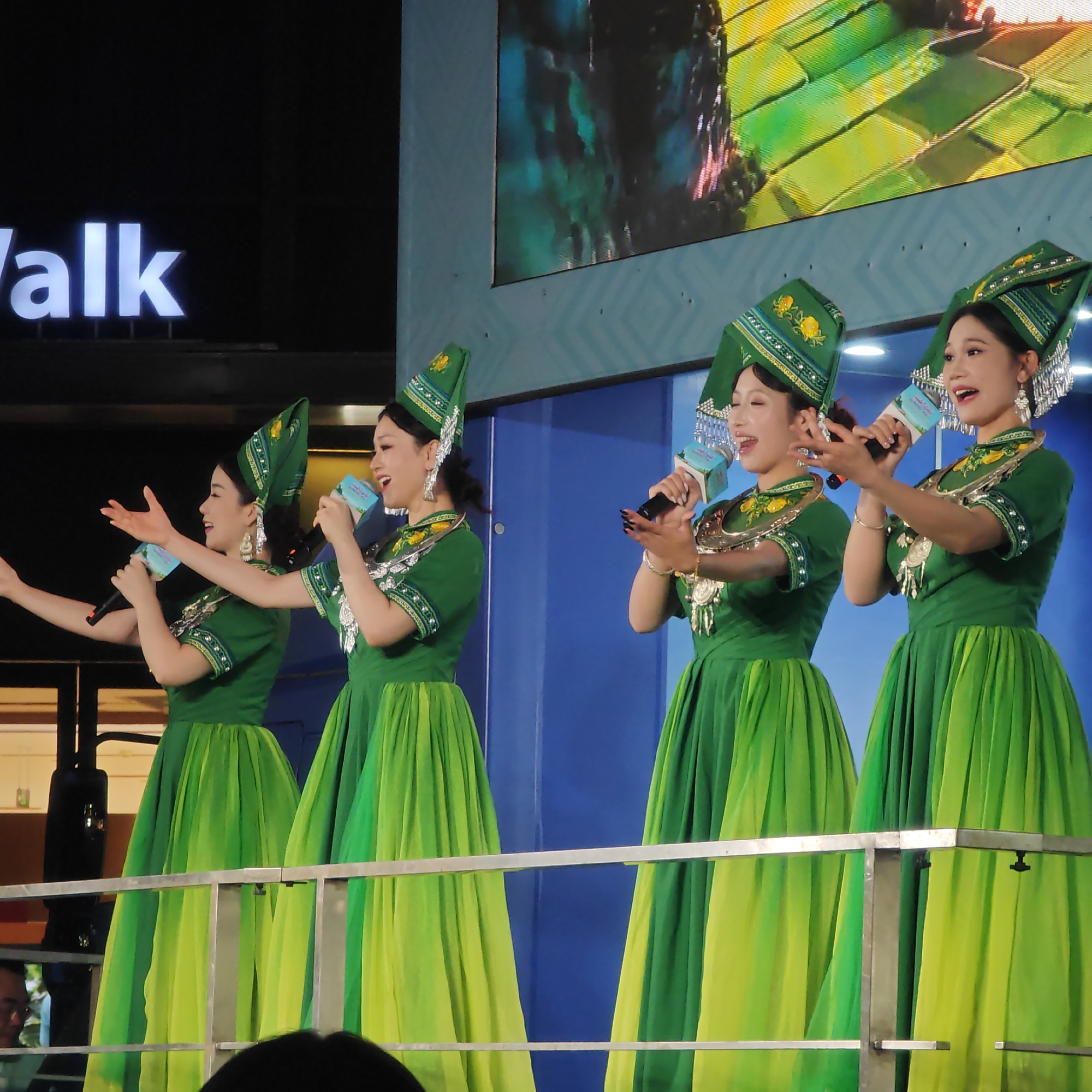 Four women are on stage, singing and gesturing with microphones. They are wearing traditional green and yellow dresses and green hats with silver tassels. Behind them is a large screen showing images of fields, and to the left is a sign that reads "Walk." The women are standing behind a silver railing. The scene is located in Yuzhong District, China.