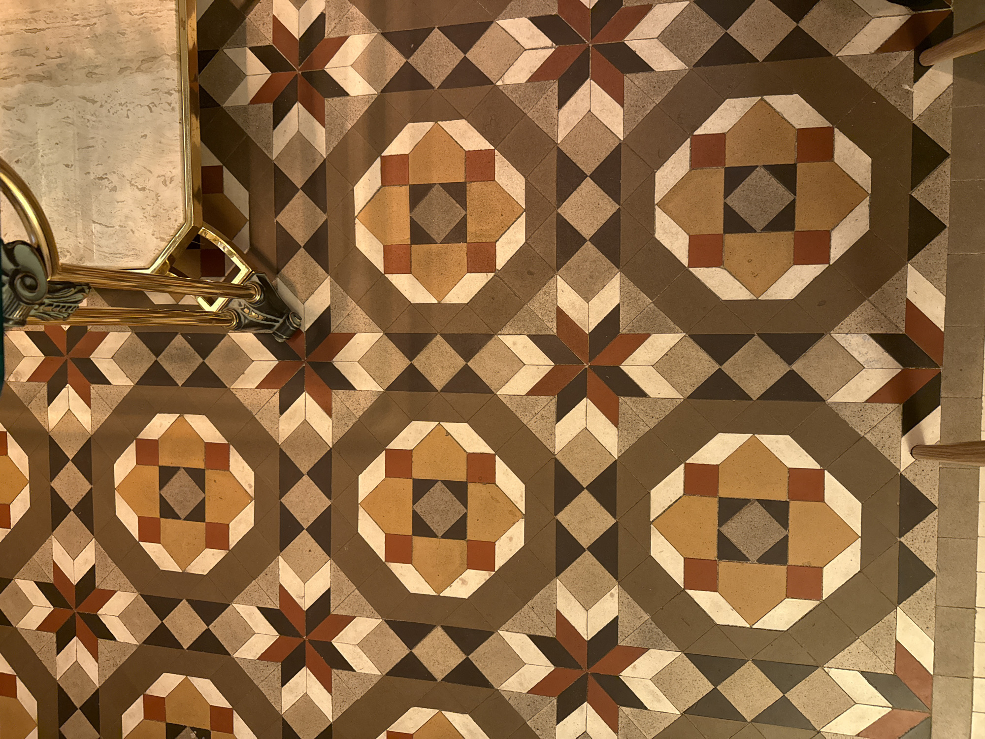 The image shows a patterned tile floor. The pattern consists of octagonal shapes interspersed with starburst designs. The colors are primarily earth tones: shades of brown, tan, white, and some brick red. A portion of a gold-colored fixture, possibly part of a furniture item or architectural detail, is visible on the left side of the image. Based on the flooring style, it is likely located in Barcelona, Spain.