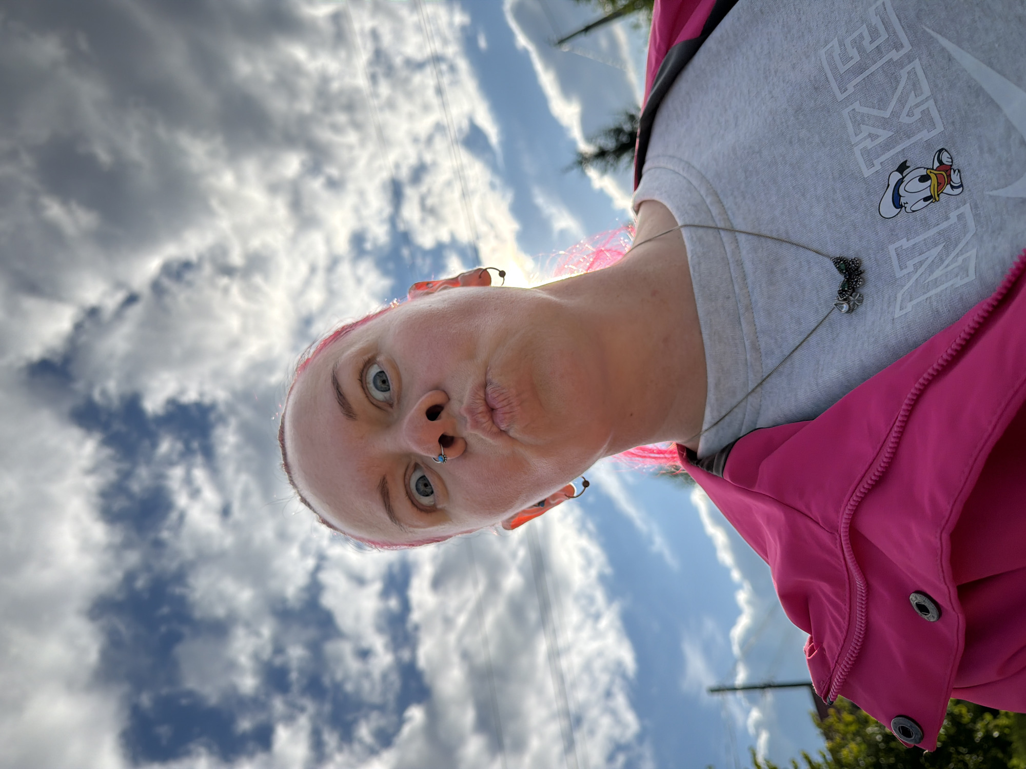 A woman with bright pink hair makes a funny face, puckering her lips and widening her eyes directly at the camera in a close-up, low-angle selfie. She is wearing a bright pink jacket over a gray sweatshirt that features a Nike logo and a cartoon image of Donald Duck. A silver necklace with a butterfly pendant hangs around her neck. The background is a cloudy sky with patches of blue, suggesting an outdoor setting. The image captures a moment of playful expression against a natural backdrop.