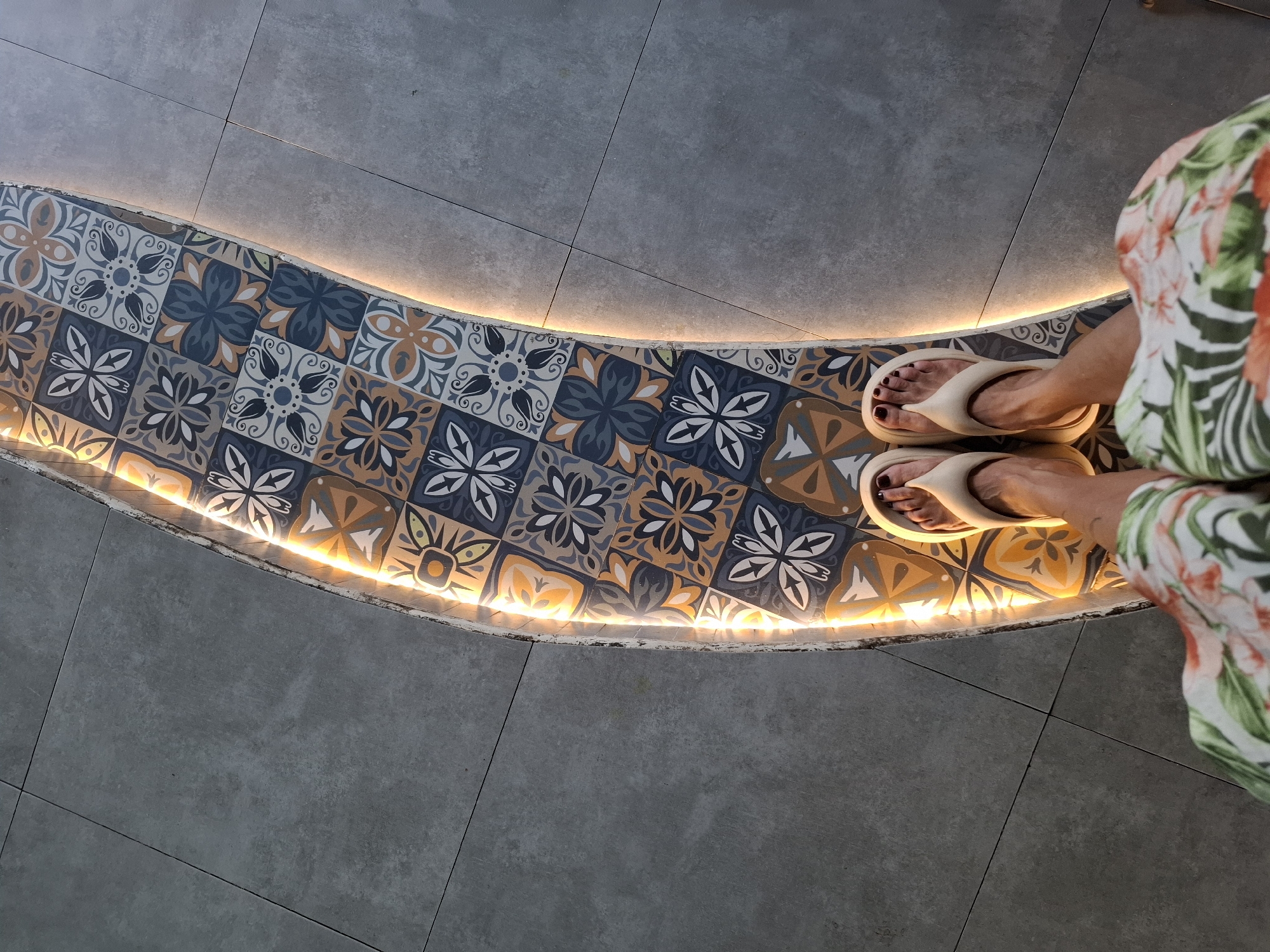 An overhead, eye-level shot shows a person's feet wearing beige flip-flops standing on a curved bench with colorful patterned tiles and a bright LED strip light running along its edge. The bench is situated on a gray tiled floor in a location in Nha Trang Ward, Vietnam. The person is wearing a white skirt with a floral print. The lighting suggests it may be indoors or a shaded outdoor area.