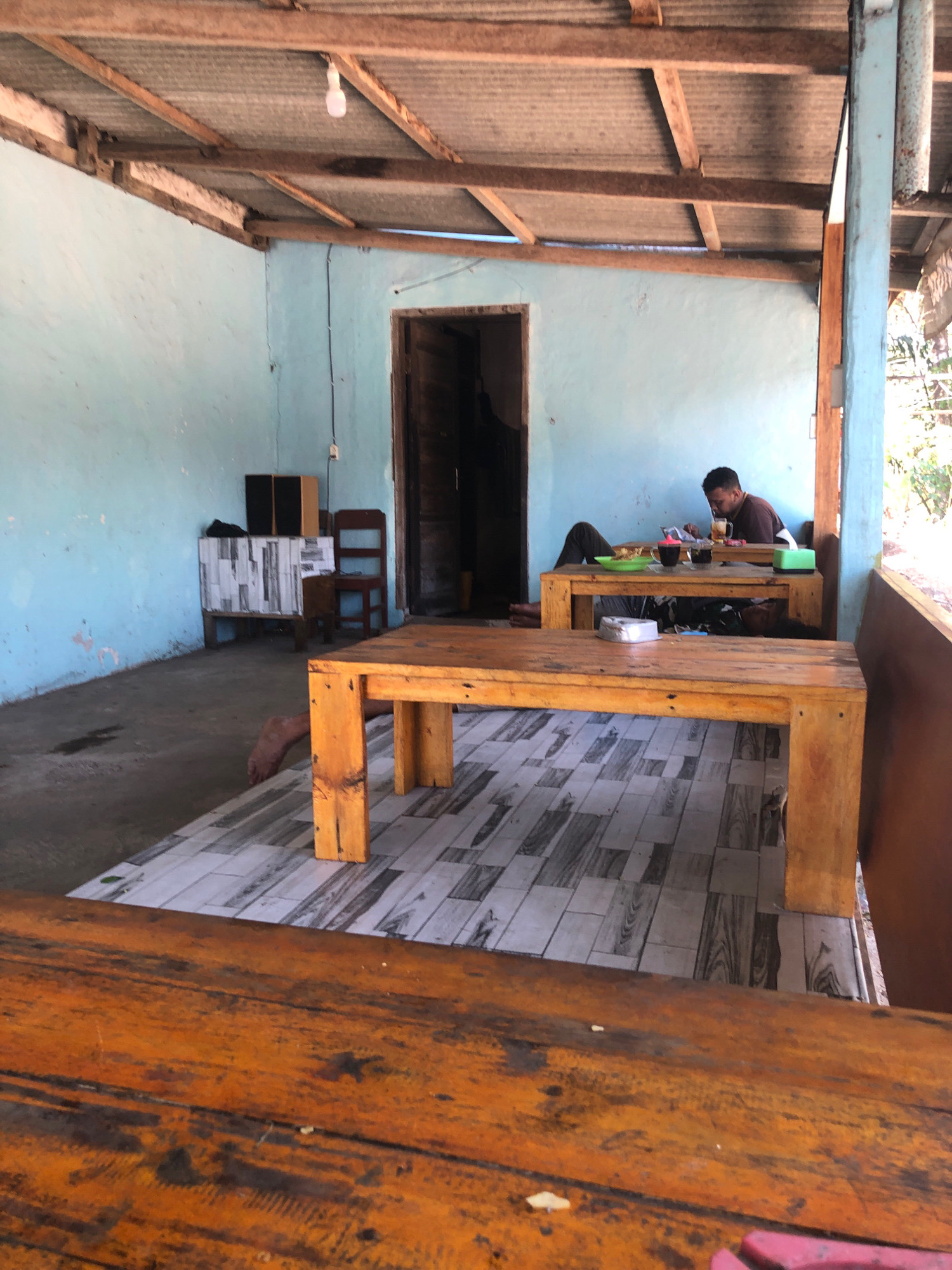 The image depicts an outdoor seating area of a casual eatery or cafe, likely in Wareng, Indonesia, given the context. The scene is characterized by rustic wooden furniture and simple construction. The ceiling is made of corrugated metal supported by wooden beams, and the walls are painted a pale blue.
In the background, a man is seated at a wooden table, appearing to be eating or drinking. He is looking down at the table, and a meal or snacks are visible in front of him. Another table, similar in construction, is positioned in front of the one the man is using. A second person's legs are visible extending from beneath this second table, suggesting they are lying down or resting there.
The floor is tiled with a pattern resembling wood planks. To the left, there's a small cabinet with two speakers on top, and a wooden chair is nearby. The overall atmosphere suggests a relaxed, informal setting, possibly a roadside stop or a local gathering place. The lighting is natural, with some shadows indicating daylight.