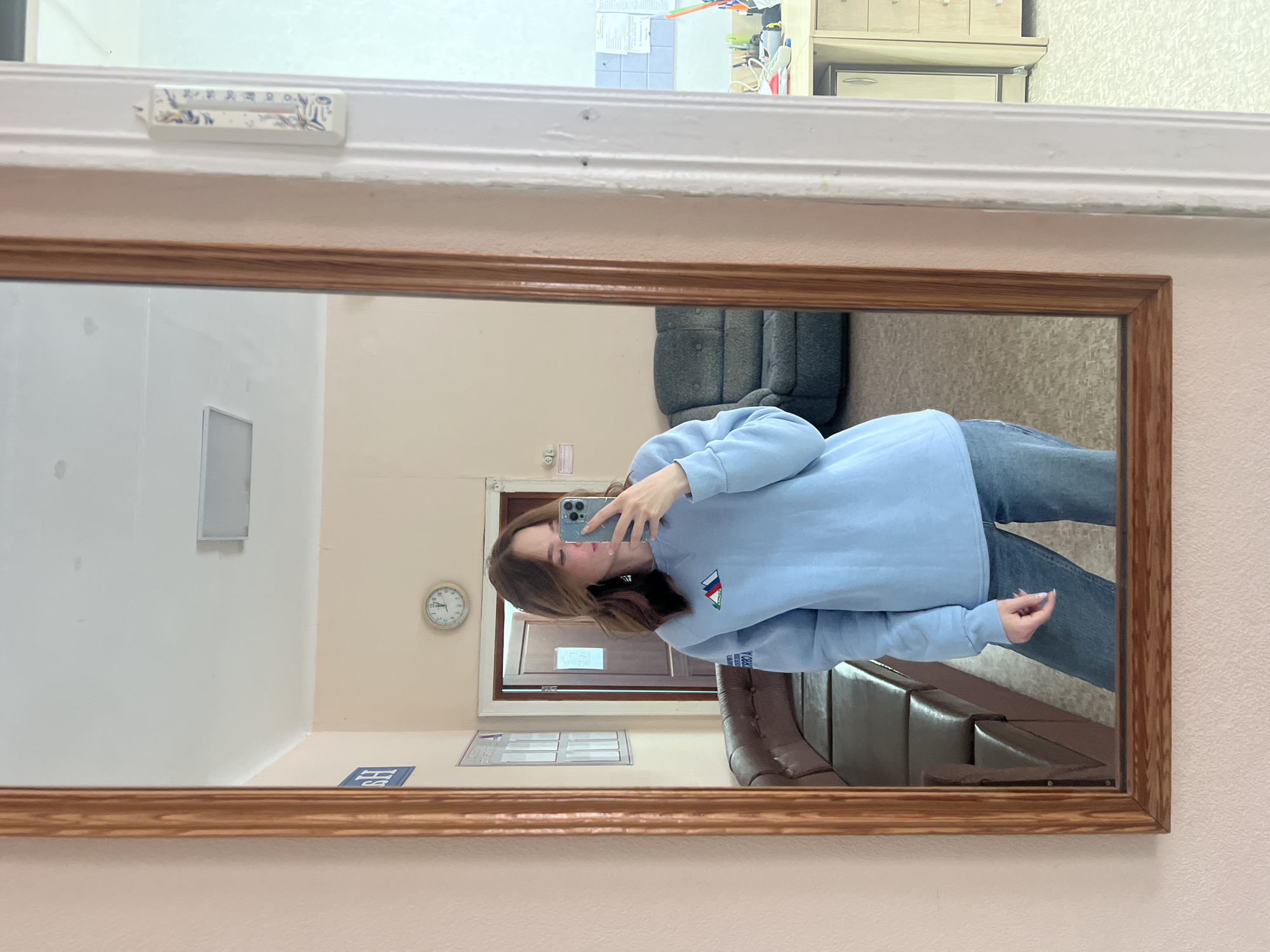 A woman is taking a selfie in a mirror. The mirror has a wood frame and is mounted on a pink wall. The woman is wearing a light blue sweatshirt and blue jeans. She is holding a blue iPhone in her left hand. The reflection in the mirror shows a room with a beige and white wall, a gray sofa, a brown leather sofa, a wall clock, and a door. A sticker with blue graphics is affixed to the wall above the mirror. Based on the visual cues of Cyrillic script and Russian imagery, the location appears to be within Russia. The presence of a logo or flag similar to the Russian flag on the woman's sweatshirt strengthens this assertion. Without more specific markers in the frame, the city of Chaikovsky cannot be confirmed; the description stops at Russian provenance.