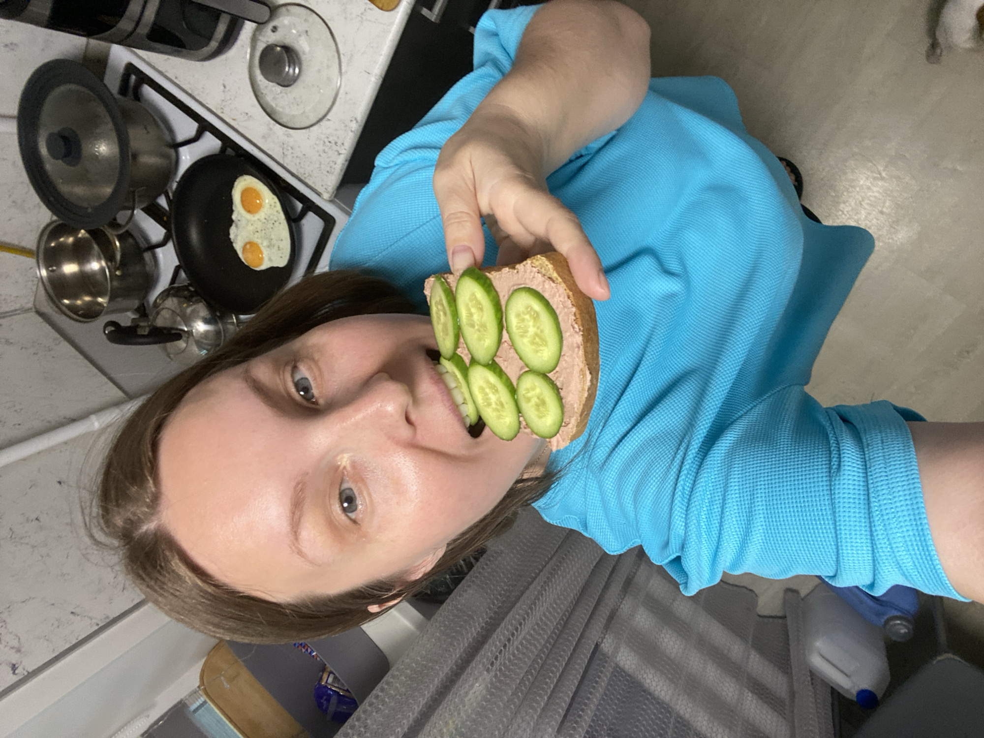 A woman with fair skin and light brown hair is holding a sandwich with liverwurst and cucumber slices towards her mouth. She is wearing a blue shirt with a waffle pattern. Behind her, a stove with a frying pan containing two fried eggs is visible, along with pots. The location appears to be in a kitchen. The image does not provide enough information to confirm the exact location as Noviy, Russia.