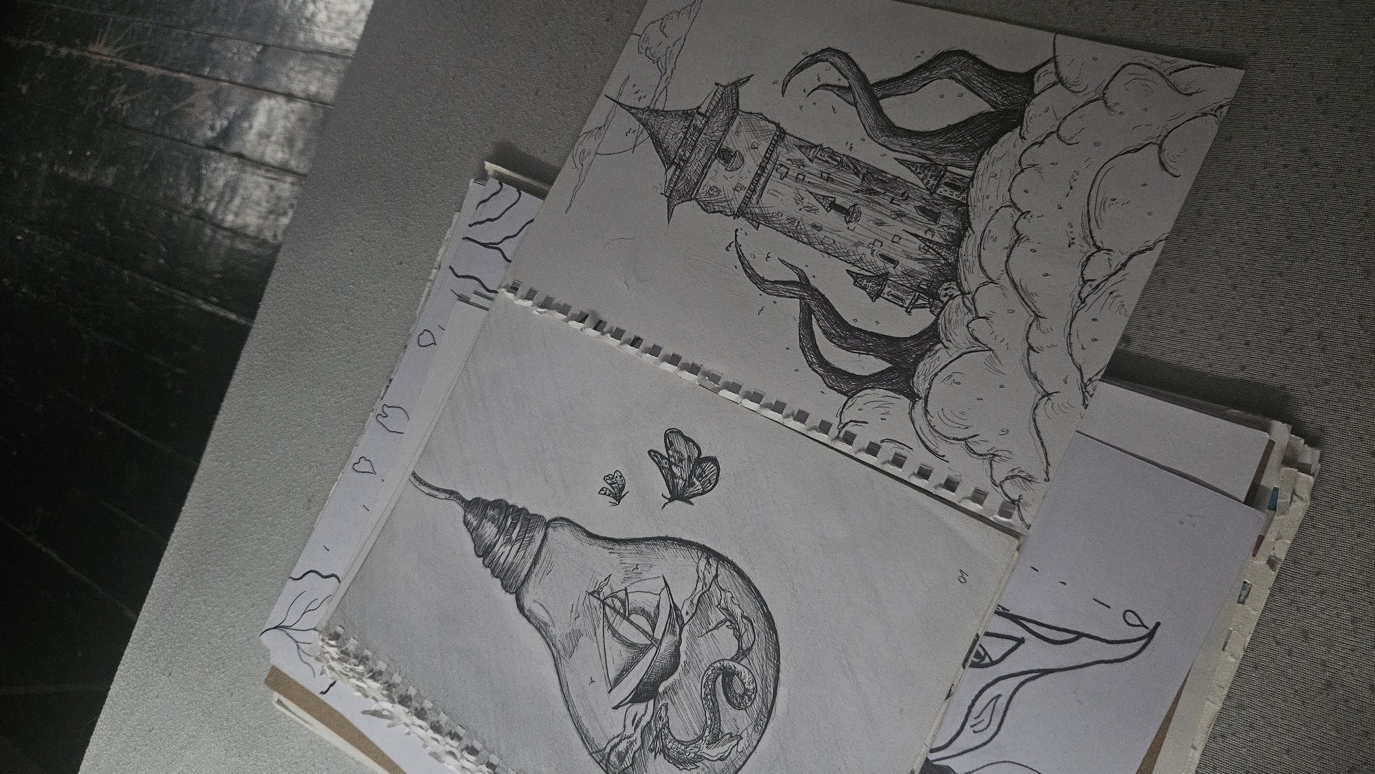 This is a close-up, high-angle shot of two ink drawings on a table. The drawings are on paper with a spiral binding along the left edge, and the paper is slightly torn and ragged. The paper with the drawing on the left depicts a lightbulb containing a sailing ship, a sea serpent, and two moths outside the bulb. The paper with the drawing on the right shows a tall, narrow tower with a conical roof, surrounded by what appear to be tentacles emerging from clouds. Both drawings are in black and white and appear to be unfinished. A third drawing, partially visible at the bottom left, features an outline of a face with elongated features. The background consists of a textured gray surface and a dark, wooden surface that appears to be flooring.