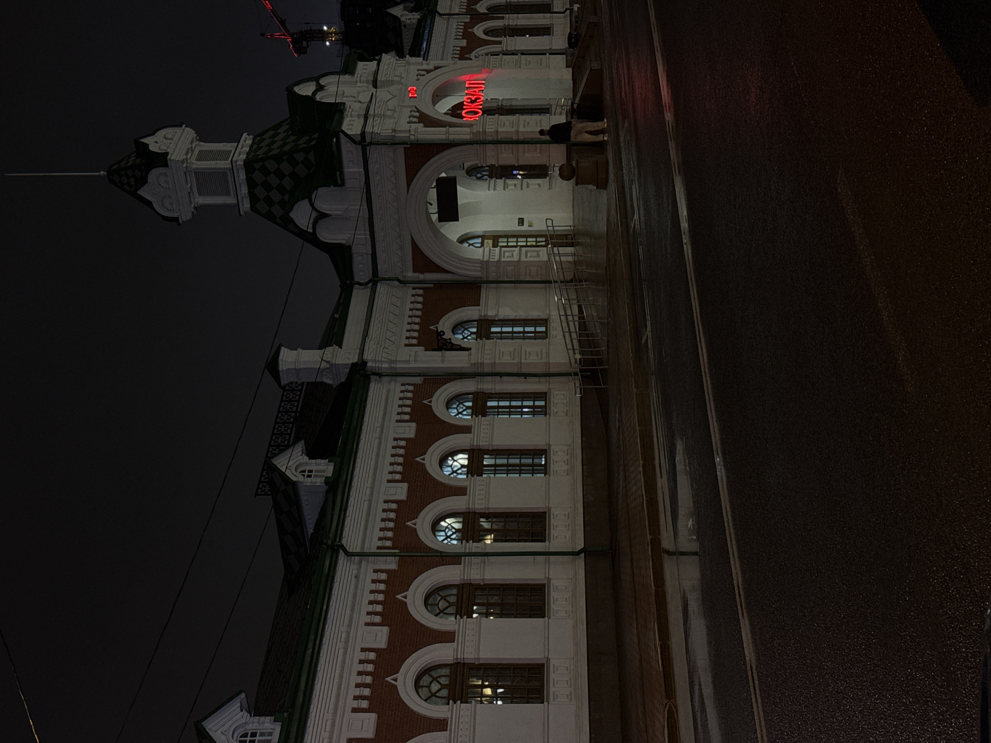 The image depicts a grand, ornate building at night, viewed from a slightly low angle across a wet street. The building is predominantly white with sections of reddish-brown brickwork, adorned with intricate white decorative trim, cornices, and pilasters. It features numerous arched windows, some circular and some rectangular, through which interior lights are visible, casting a warm glow.
A prominent architectural feature is a multi-tiered tower located on the left side of the building (when oriented upright), topped with a dark, possibly checkered or diamond-patterned roof and a slender spire. Towards the center of the building, a large archway marks a main entrance. Above this arch, a bright red neon sign spells out "ВОКЗАЛ" (VOKZAL), which is Russian for "station" or "terminal."
Underneath the main archway, a couple of dark, silhouetted figures are faintly visible, suggesting people either entering, exiting, or waiting near the entrance. The street in the foreground and to the right is dark and appears wet, possibly from recent rain, reflecting faint light sources from the building or distant unseen lights. The sky above is uniformly dark, confirming the nighttime setting.
The overall scene conveys a quiet, somewhat majestic atmosphere, characteristic of a historic public building late at night. Given the text "ВОКЗАЛ" and the architectural style, it is consistent with a railway station, specifically the Perm-2 railway station in Perm, Russia, based on the provided location context.
