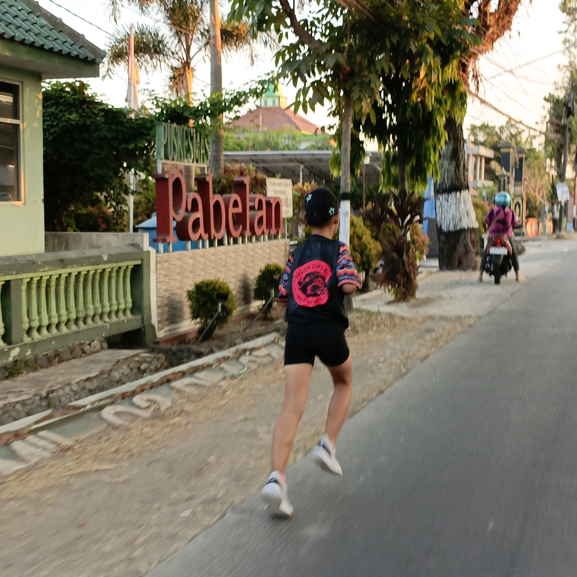 In Pabelan, Indonesia, at approximately dusk, a young person with dark hair, wearing a black t-shirt with a red circular graphic and multi-colored sleeves, and black shorts, runs along a paved road. Their white athletic shoes are visible as they run. Behind them, a woman on a motorcycle, wearing a purple jacket and a blue helmet, travels away from the viewer. The environment includes a light green building with a tiled roof on the left, a green railing, and a large sign with red lettering that reads "Pabelan." Several trees are present, some with white paint on their trunks, and power lines are visible overhead. The sky is a pale orange, indicating the end of daylight.