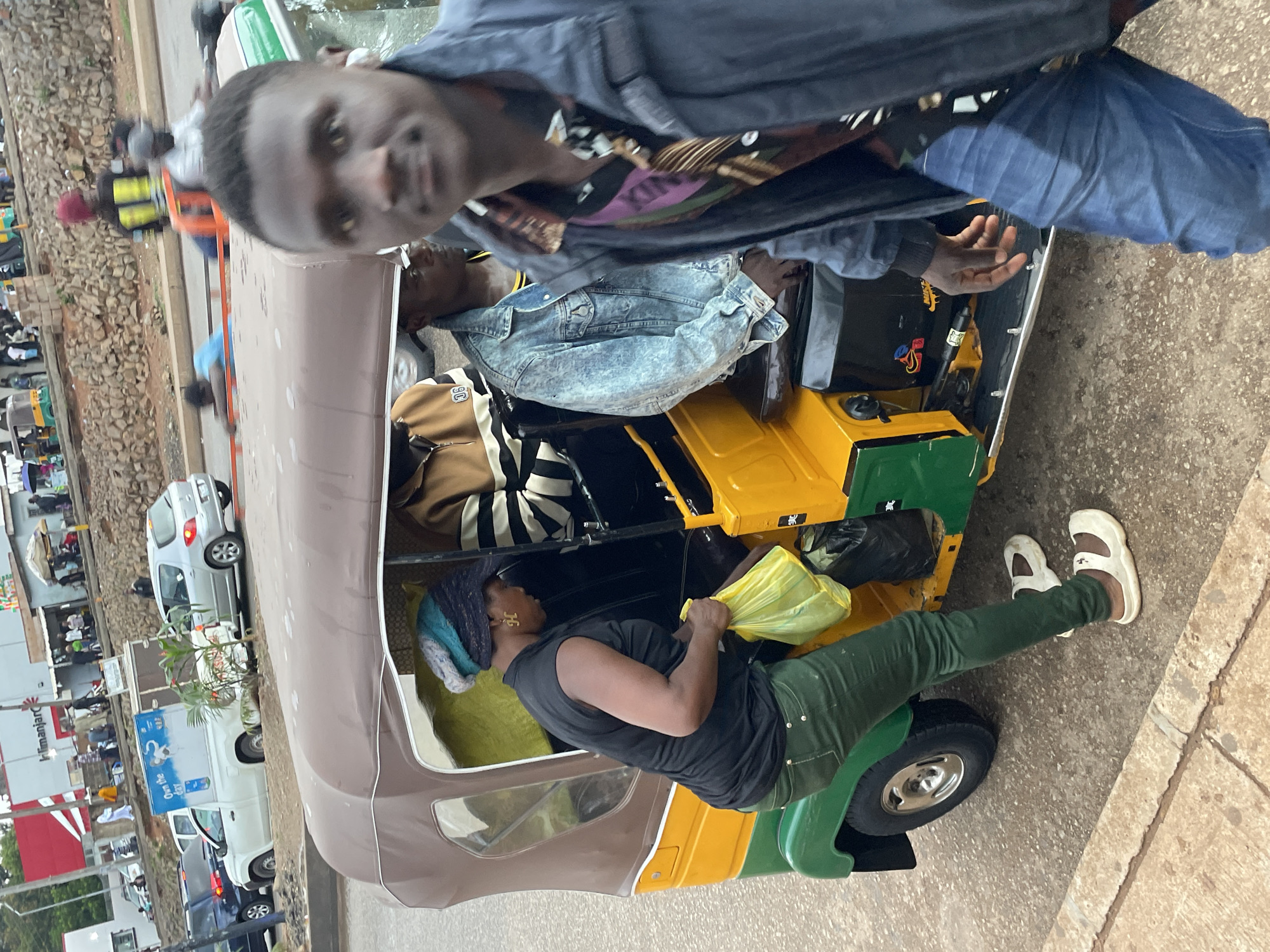In a street scene in Jos, Nigeria, a man in the foreground walks towards the camera, blurred and partially obscuring the view of a colorful auto-rickshaw. The auto-rickshaw, predominantly yellow and green with a brown canvas top, is occupied by several individuals. A woman in a black sleeveless top and green pants is standing next to the open door of the auto-rickshaw, holding a yellow plastic bag. Another person wearing a denim jacket is seated inside the auto-rickshaw. The background shows more traffic, including a white pickup truck with a blue graphic on its side and other vehicles, as well as buildings and a rocky embankment lined with trees. The street is paved, and a sidewalk is visible in the foreground.