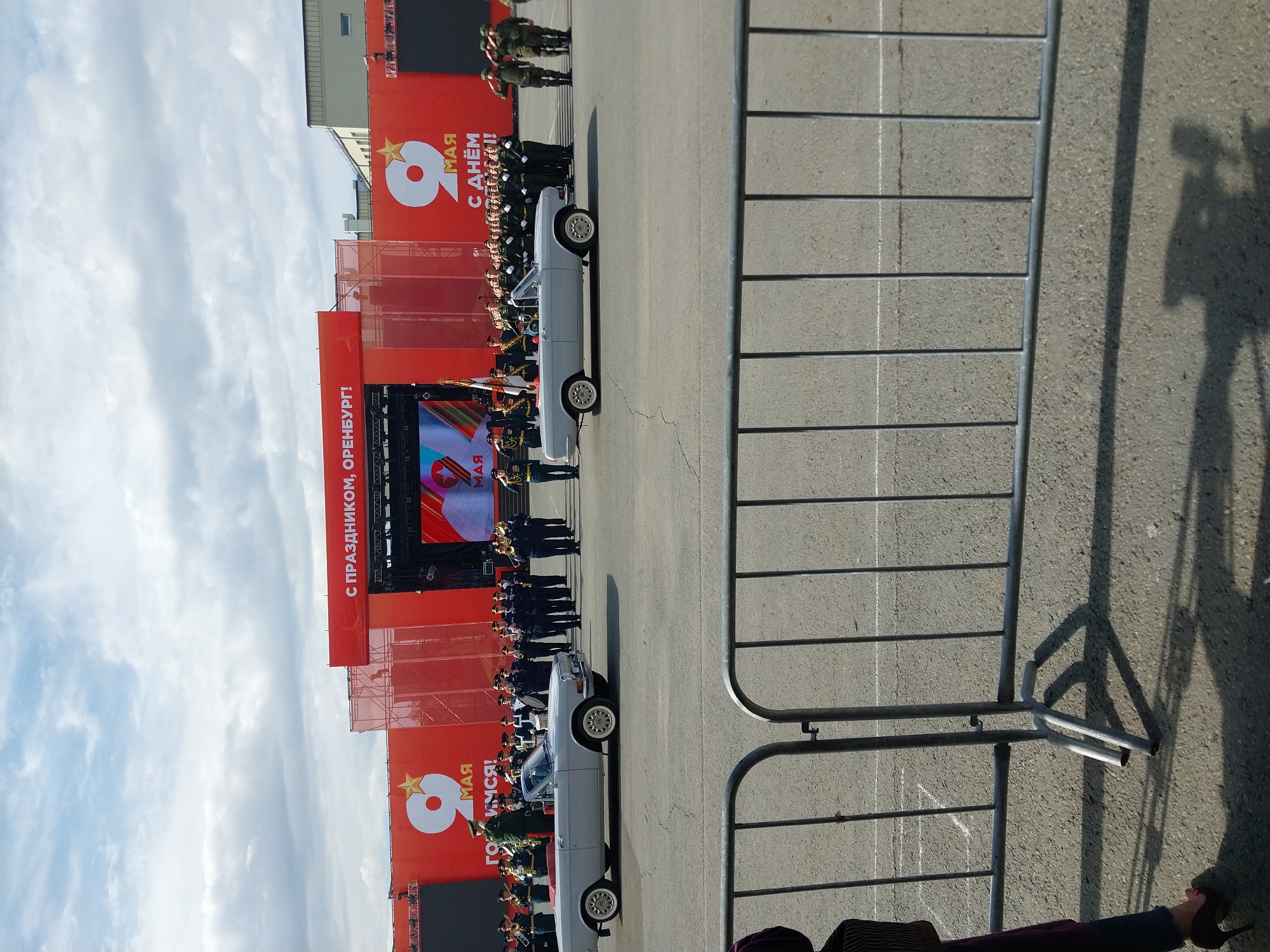 The image depicts a Victory Day parade in Orenburg, Russia. Red banners with the number "9" and the words "MAY" and "WITH THE DAY OF VICTORY!" are visible. Soldiers in dress uniform stand in formation near vintage cars. A metal barrier separates the parade from onlookers. The sky is partly cloudy.