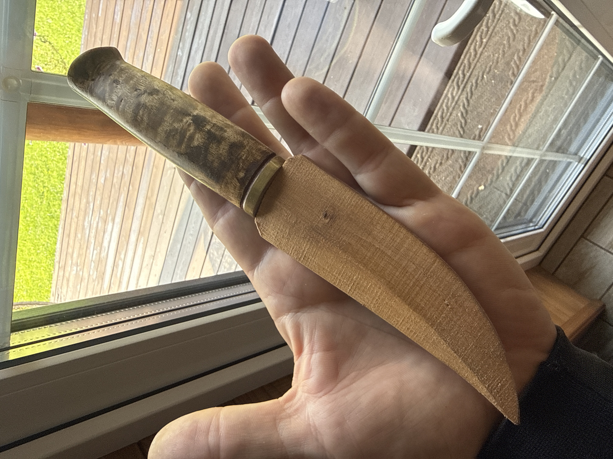 A person's hand is holding a knife with a wooden handle and a wooden blade. The knife is positioned diagonally across the palm. The background shows a wooden deck through a window with green grass and trees visible beyond. The scene appears to be outdoors, possibly in a residential area.
