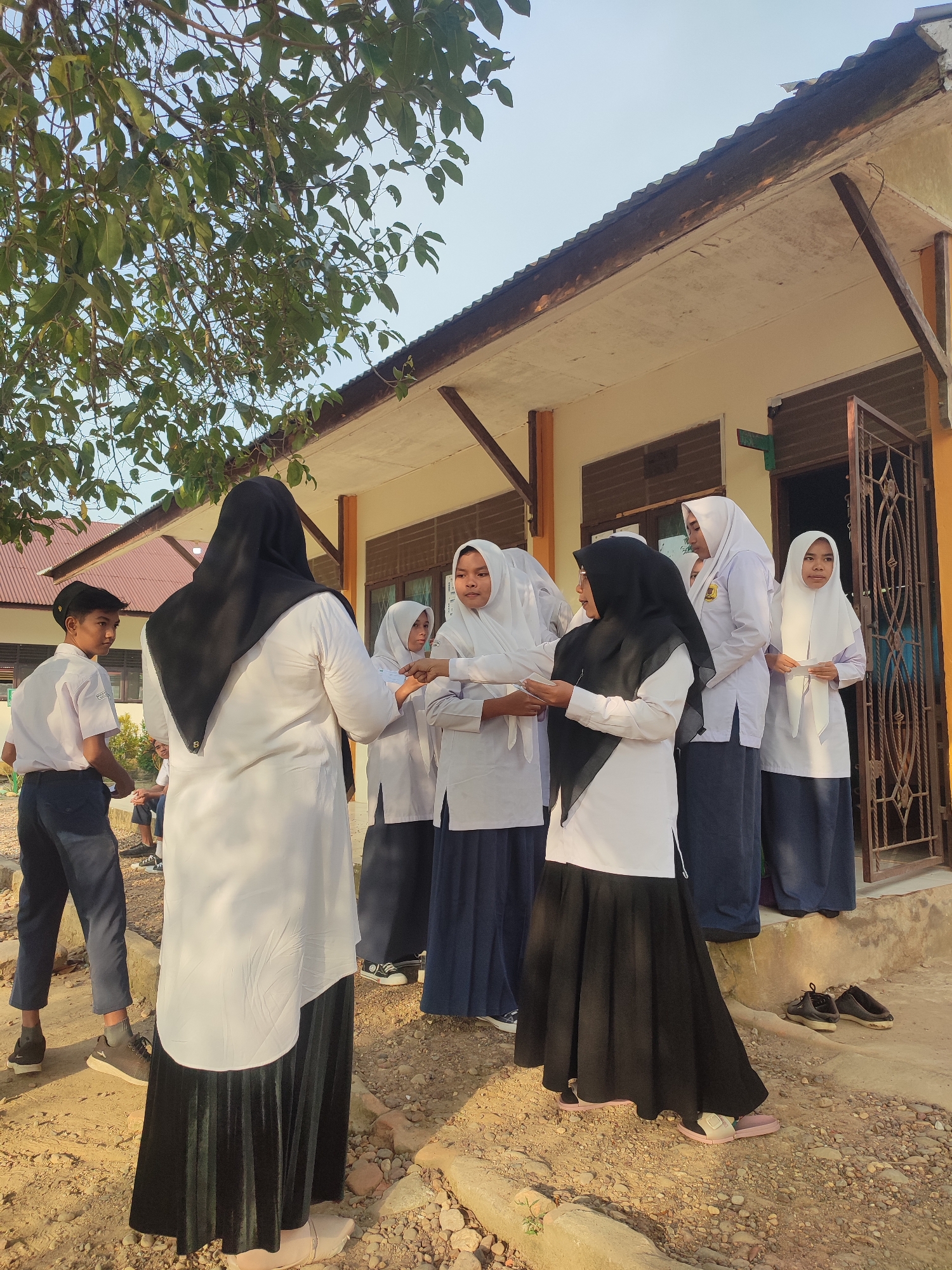 A group of Indonesian students and an adult are gathered outdoors in front of a school building. The majority of the individuals are young females wearing white long-sleeved shirts, dark skirts, and hijabs, typical school uniforms. One boy is visible on the left, also in a white shirt and dark pants. An adult woman, distinguished by her black hijab and long white dress-like top, stands in the center with her back to the camera.
The central activity involves several female students exchanging or receiving white slips of paper or folded documents from each other or the adult. Some students are standing on a raised porch area near the building's entrance, while others are on the dirt ground below.
The scene takes place outdoors on a bright, clear day, likely morning or late afternoon, as indicated by the strong natural light and some visible shadows. The setting is a simple, single-story building with a corrugated metal roof, possibly a classroom or school office. The ground is unpaved, consisting of dirt and gravel. A tree with green leaves partially covers the upper left of the frame, providing some shade. Various shoes, including sneakers and sandals, are seen on the ground near the steps. No clear, readable text is visible in the image. The overall atmosphere appears calm and routine, consistent with a school environment in Blangriek, Indonesia.