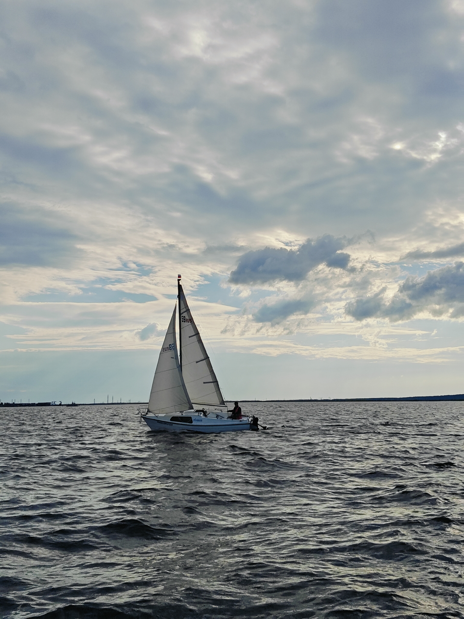 A white sailboat with a red, white, and blue flag on its mast sails on dark, choppy water under a cloudy sky. The boat has two sails deployed and appears to be moving from left to right. There are two people visible on the boat; one is standing near the helm and the other is seated closer to the bow. The background shows a distant shoreline with industrial structures and power lines. The scene is set on the Kama River in Chaykovsky, Russia, a city known for its hydroelectric power station and its location on the river. The lighting suggests it may be late afternoon or early evening.