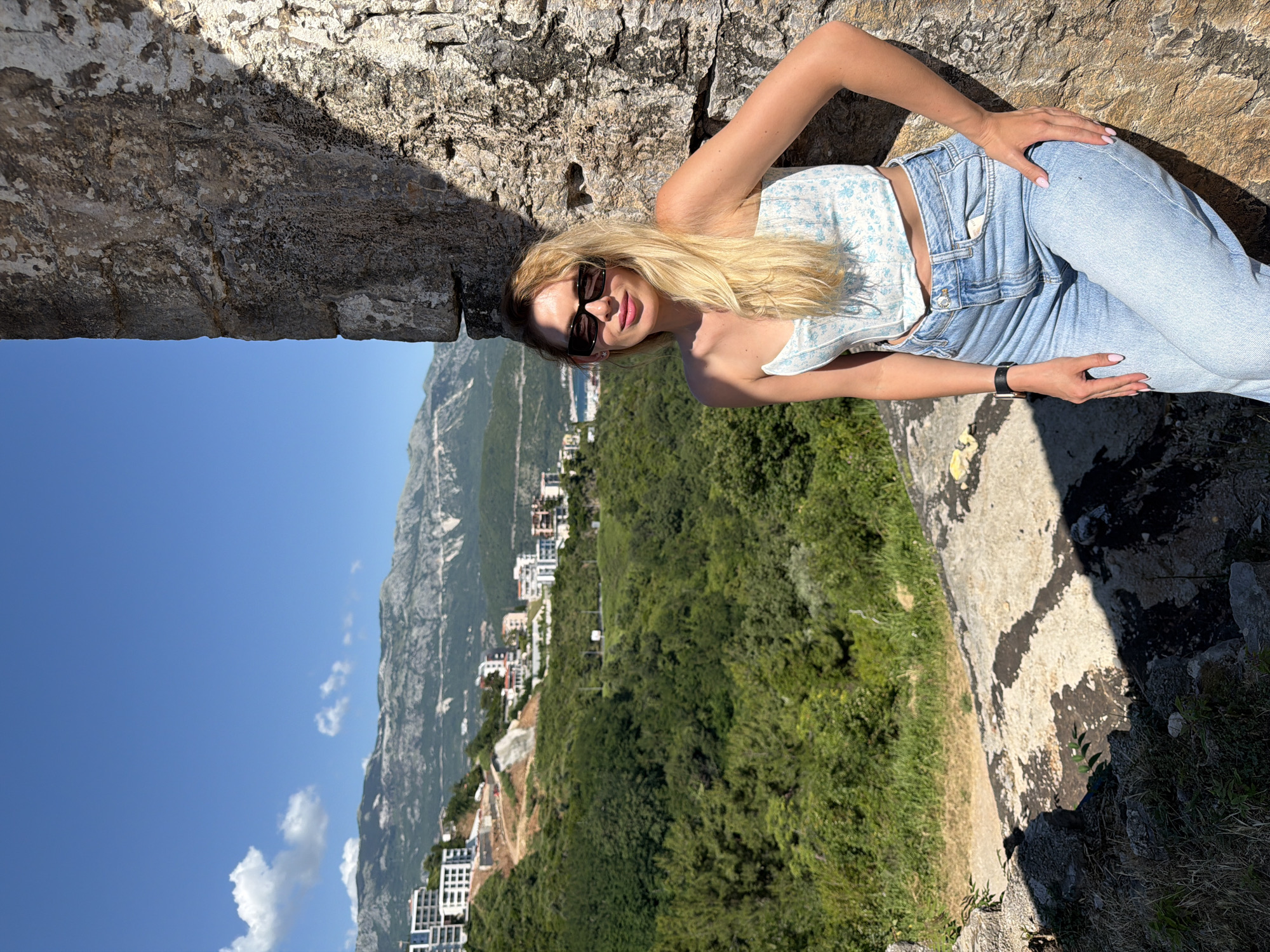 A young adult female with long blonde hair, wearing black sunglasses, a light blue and white floral strapless top, and light blue jeans, stands leaning against a rough, weathered gray stone wall. She has light pink nail polish on her fingers and a black smartwatch on her left wrist. Her left hand rests on her left thigh, and her right arm is bent, propping her against the wall. She looks directly forward with a subtle smile. The scene is brightly lit by natural sunlight, creating strong shadows on the stone wall and ground.
To the left of the woman, a panoramic view of Budva, Montenegro, unfolds under a clear blue sky with sparse white clouds. A steep, verdant hillside covered in dense green trees and shrubs descends towards the coast. Numerous modern, multi-story white residential buildings with rectangular windows are scattered across the upper and mid-slopes of the hillside. A paved road is visible winding through the greenery. In the distance, a segment of the blue Adriatic Sea is visible, with additional buildings along its shore. A prominent range of rugged, gray mountains with visible rocky patches forms the distant background horizon. The ground in the immediate foreground, to the left of the woman, consists of rocky, dusty terrain with sparse vegetation.