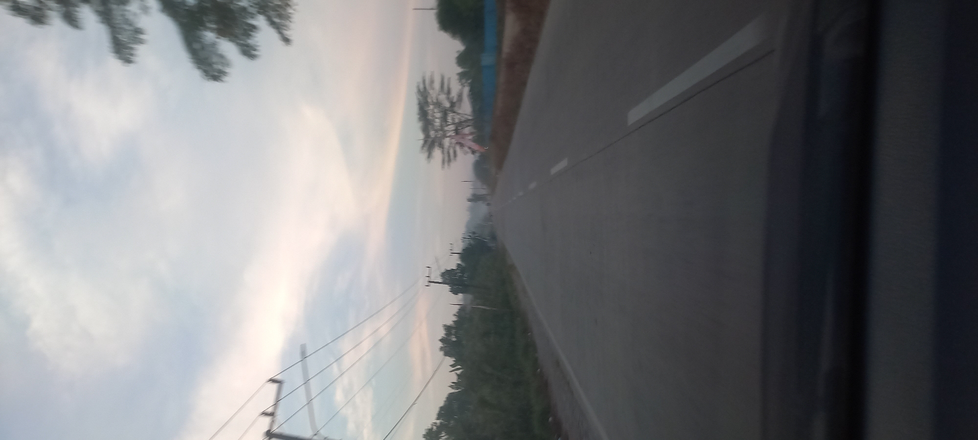 The image depicts a view from a vehicle on a road in Sepakat, Indonesia, likely during dawn or dusk. The sky is overcast with soft, pale light, suggesting low light conditions. On the left side of the road, there are utility poles with numerous wires extending across the frame. Further down the road, a line of trees and vegetation is visible. The road itself is asphalt with white lines marking the lanes. On the right side of the road, there are more trees and some fencing or walls. The foreground shows the dashboard of the vehicle from which the photo was taken. There are no visible people in the scene. The overall atmosphere is calm and slightly hazy.