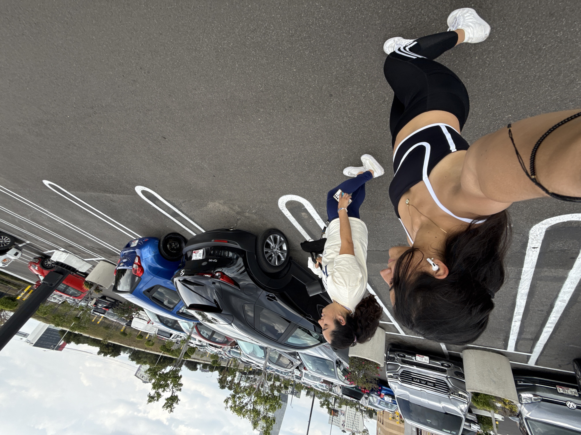 Two women are standing in a parking lot. One woman is wearing a black and white sports bra, black leggings, and white sneakers. The other woman is wearing a white shirt, blue pants, and white sneakers, holding a phone. The parking lot has many cars, including a blue hatchback and a black SUV. There are buildings and trees in the background. The location is not specified in Puebla, Mexico.