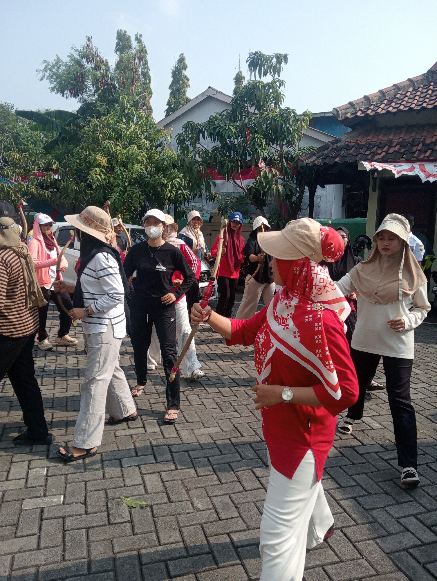 In Demak, Indonesia, a group of women are participating in an outdoor activity on a paved brick surface. The scene appears to be a street or courtyard in a residential or community area. The women, some wearing headscarves and hats, are dressed in casual attire, with some wearing long-sleeved shirts and pants. Several of the women are holding what appear to be wooden sticks, possibly for a game or exercise. One woman in the foreground is wearing a red top and white pants, and a red and white patterned scarf. She has a watch on her left wrist. Another woman in a black shirt and pants, wearing a white face mask, stands with her hands on her hips. The background shows trees, houses with tiled roofs, and a white vehicle. The lighting suggests it is daytime, with sunlight casting shadows.