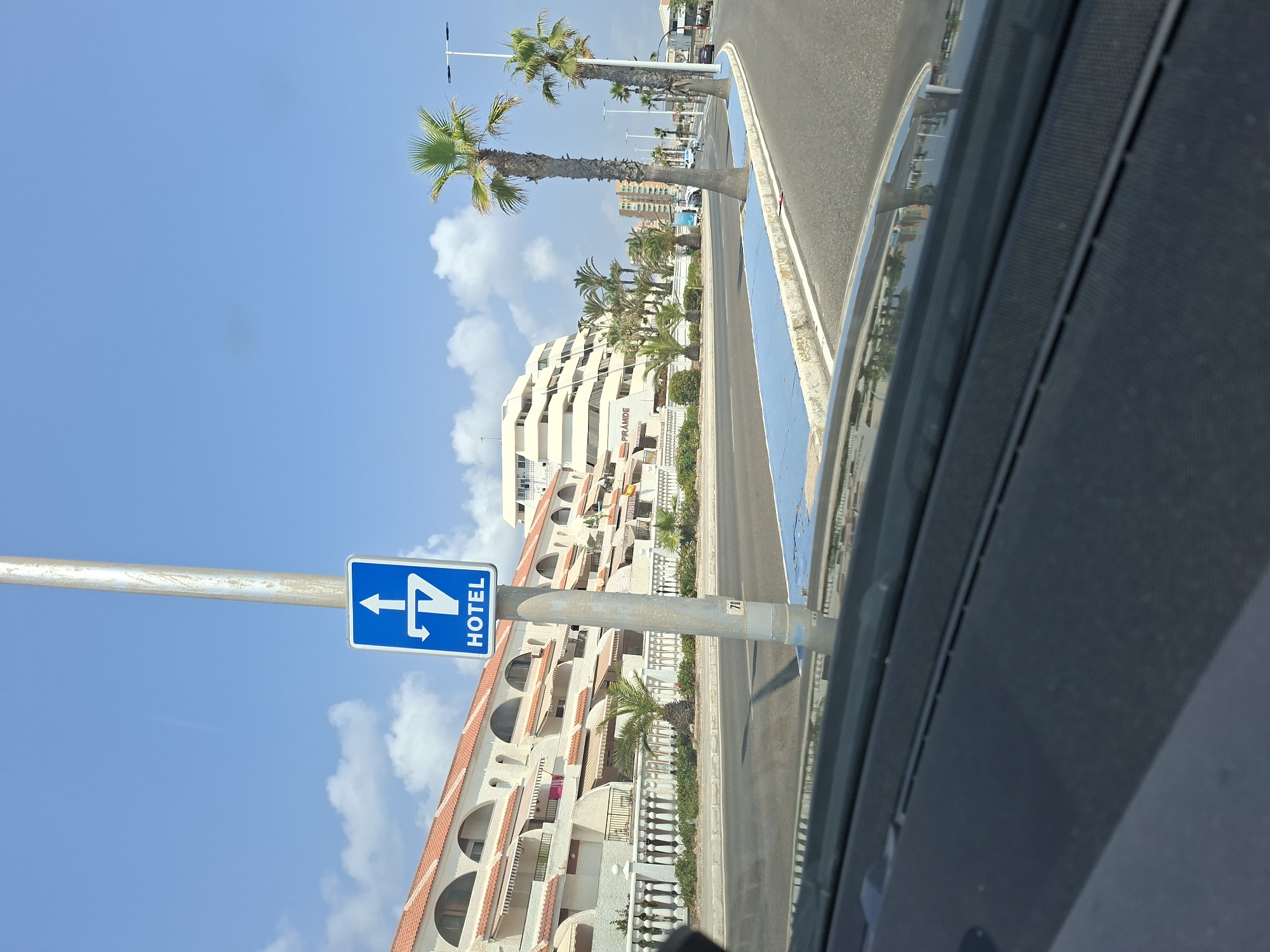 The image shows a street scene in La Manga Del Mar Menor, Spain. A blue and white sign on a metal pole indicates "HOTEL" with a directional arrow pointing straight ahead and a left turn. The buildings in the background appear to be apartments or hotels with white facades and orange tiled roofs. Palm trees line the street, and the sky is blue with scattered clouds. The perspective suggests the photo was taken from inside a vehicle, as the dashboard is visible in the foreground.