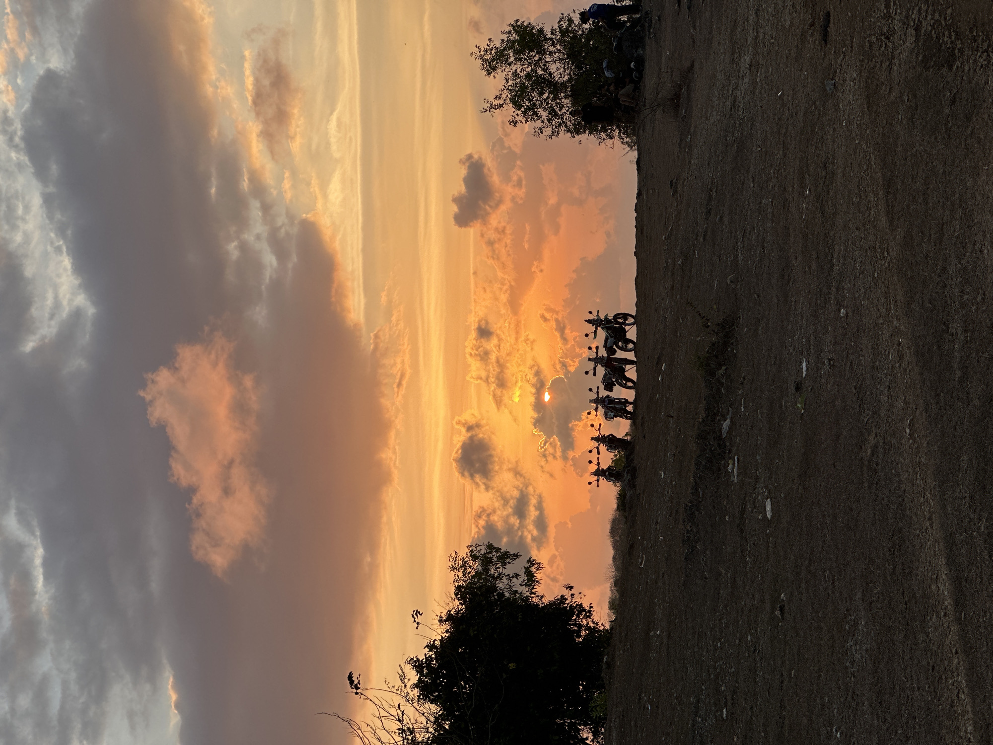 The image depicts a silhouetted scene at sunset on a hilltop, likely in Kuta Selatan, Indonesia, given the tropical vegetation. In the foreground, the ground is dry and covered with sparse grass and scattered debris. A line of four motorcycles is parked on the crest of the hill, silhouetted against the vibrant sky. To the left, a large, dark tree dominates the silhouette, its branches bare. To the right, another tree is visible, with a small group of people gathered underneath it, also in silhouette. The sky is filled with dramatic clouds, painted in hues of orange, pink, and yellow, with the setting sun peeking through a gap in the clouds, casting a warm glow. The overall impression is one of a serene, natural landscape at dusk.