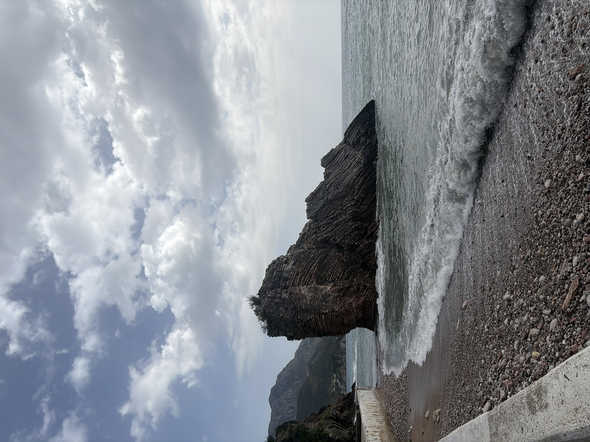 The image depicts a rocky coastline. A large, stratified rock formation dominates the scene, situated at the edge of the water. Waves are crashing against the pebble beach. The sky is overcast with a mix of white and gray clouds. In the background, a mountainous landscape is visible. The image is oriented vertically. The location is Unknown, Unknown.