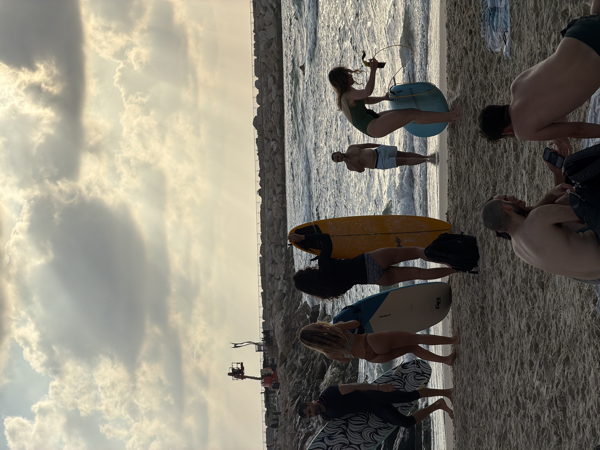 In Tel-Aviv, Israel, on a sandy beach, a group of people are gathered near the water. A man in a black wetsuit stands on the left, holding a surfboard. Next to him, two women in bikinis are also holding surfboards, one blue and one yellow. A man in swimming trunks stands in the water, and a woman in a green swimsuit is seen interacting with a blue surfboard. Further down the beach, two men are seated on the sand, one looking at his phone. The ocean is visible with waves, and a stone jetty extends into the sea in the background. The sky is filled with dramatic clouds.