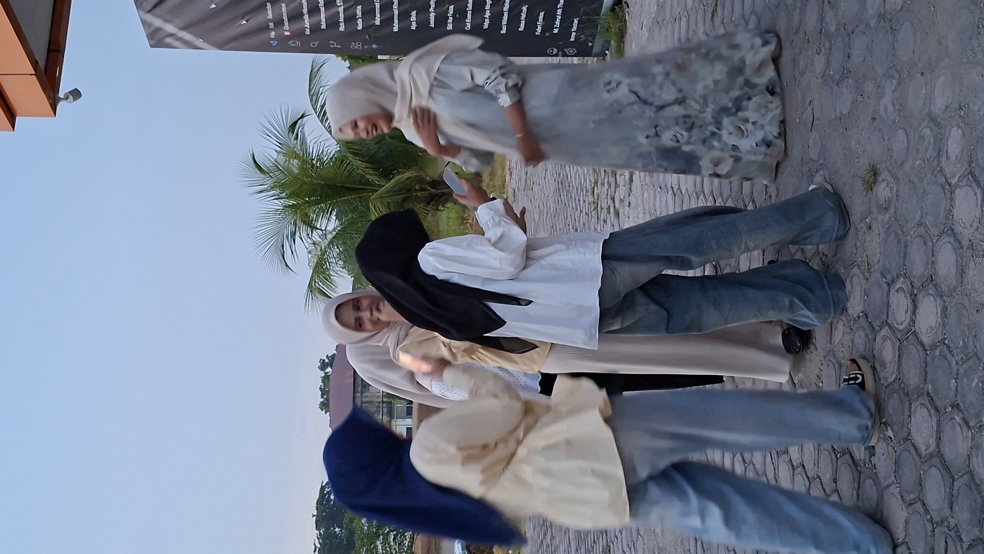 Four young women in hijabs are gathered outdoors, possibly in front of a building with an orange overhang, in Lhokseumawe, Indonesia. The woman on the far left wears a dark blue hijab and a light yellow blouse, with light blue pants. The woman next to her, wearing a light beige hijab and dress, is partially obscured. The woman in the center has a black hijab and a white long-sleeved shirt with denim pants. The woman on the far right wears a light gray hijab and a patterned dress. The ground is paved with hexagonal stones. A palm tree is visible in the background, along with a building with a textured facade featuring text. The sky is overcast. The women appear to be interacting with each other, with two of them looking at a smartphone.