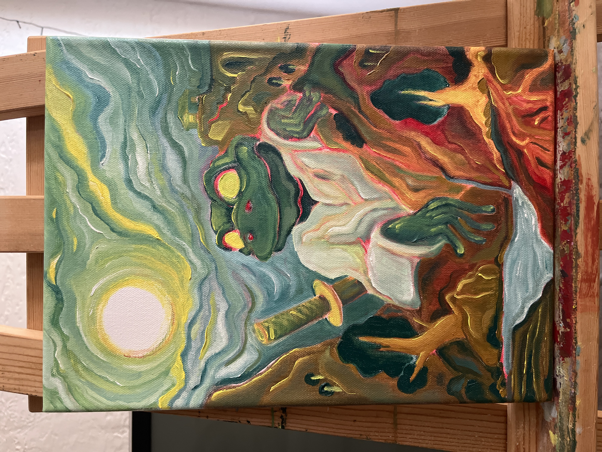 The image shows a painting resting on a wooden easel. The painting depicts a green-skinned figure, possibly a frog-like humanoid, in a white robe, wielding a sword. The figure has large yellow eyes with red pupils. The background features swirling patterns of green, yellow, and blue on the left side, suggesting a celestial or atmospheric scene with a bright, circular light source. The right side of the painting transitions into earthy tones of brown, red, and gold, resembling a forested area or terrain. The painting is on a stretched canvas, and the easel is made of light-colored wood.
