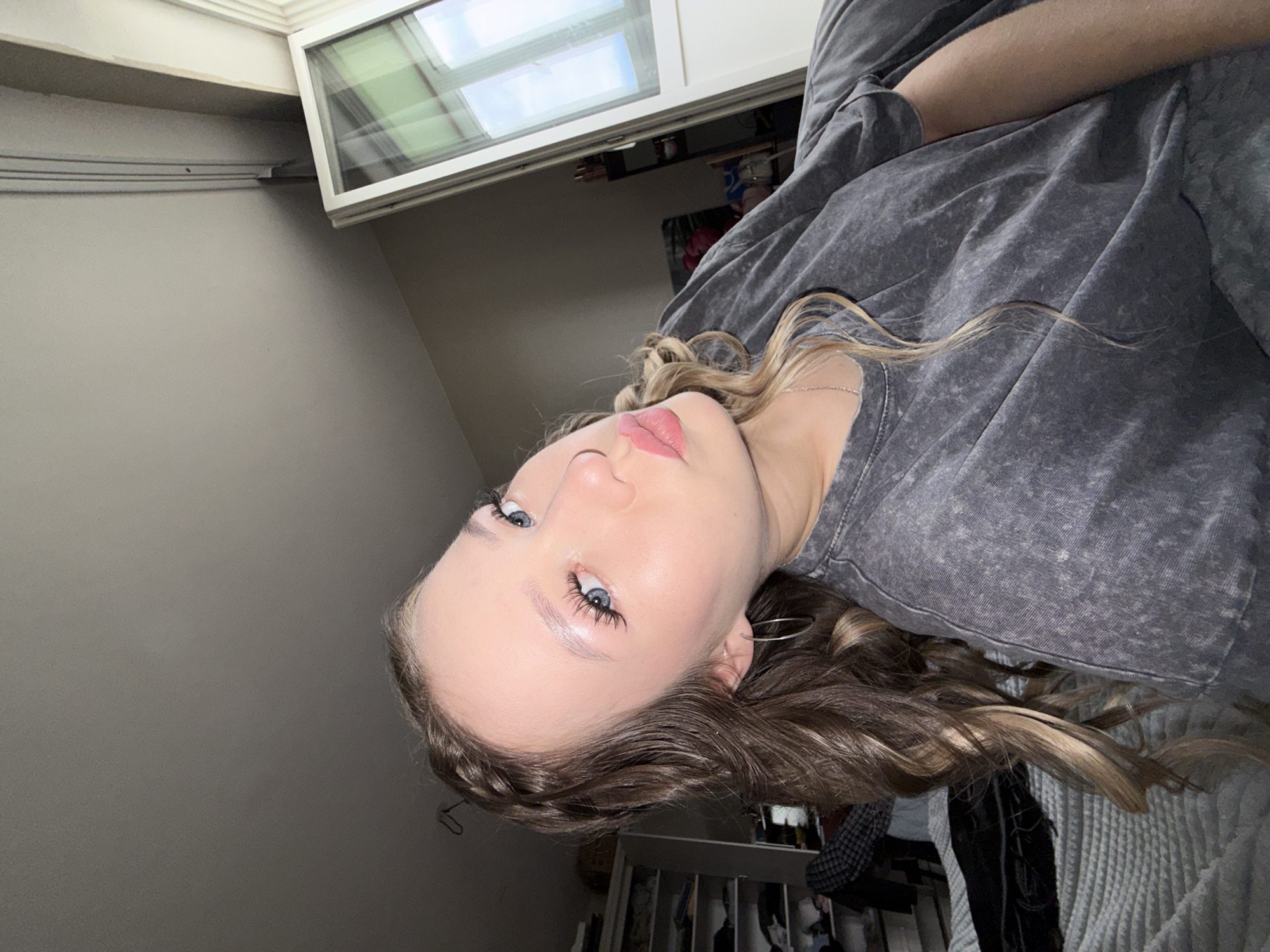 The image shows a young woman with blonde hair styled in loose waves and a braid on the side. She is wearing a grey t-shirt and has on makeup, including noticeable false eyelashes and pink lipstick. She is leaning forward, with her head tilted and her lips pursed in a kiss pose, looking towards the camera. Her right eye is prominently in focus, showing her blue iris and long lashes. She has a silver necklace and a hoop earring visible.
The background suggests an indoor setting, likely a bedroom. There's a white bed with a textured comforter in the foreground. To the left, a white wardrobe or shelving unit filled with clothes is partially visible. To the right, a large window with white framing is present, revealing reflections of the sky and possibly greenery outside. The room appears to be lit by artificial light, possibly from above, as indicated by the shadows and the bright lighting on the woman's face. The overall impression is of a casual, personal space.
The request for information regarding the "location in the city of Chaikovsky, Russia" cannot be fulfilled as there are no identifying landmarks or contextual clues within the image that would allow for its specific geolocation. The scene depicted is a generic indoor setting that could be located anywhere.