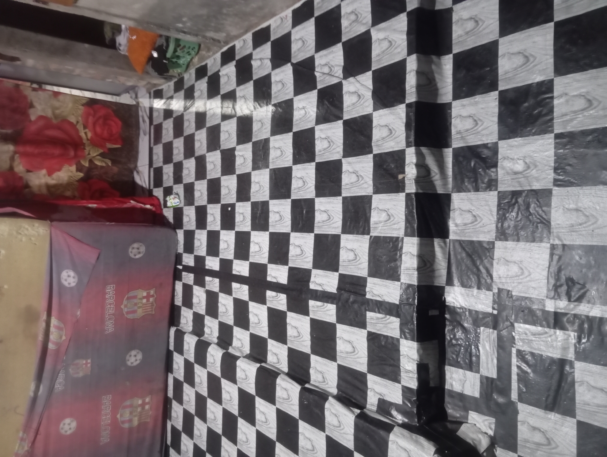 The image shows a tiled floor with a black and white checkered pattern. The tiles appear to be made of plastic or linoleum, and have a wood-grain texture printed on them. The floor is in a dimly lit room, possibly a bedroom or living area. In the background, there is a red and black bedspread with a Barcelona FC logo and soccer balls printed on it. There is also a curtain with a floral pattern of red roses. To the right of the image, there are some stacked orange and green objects, possibly laundry baskets. The overall impression is one of a cluttered and somewhat worn living space. The image is taken from a low angle, looking across the floor. The location is identified as Dente Teladas, Indonesia, although no specific landmarks or details in the image confirm this location.
