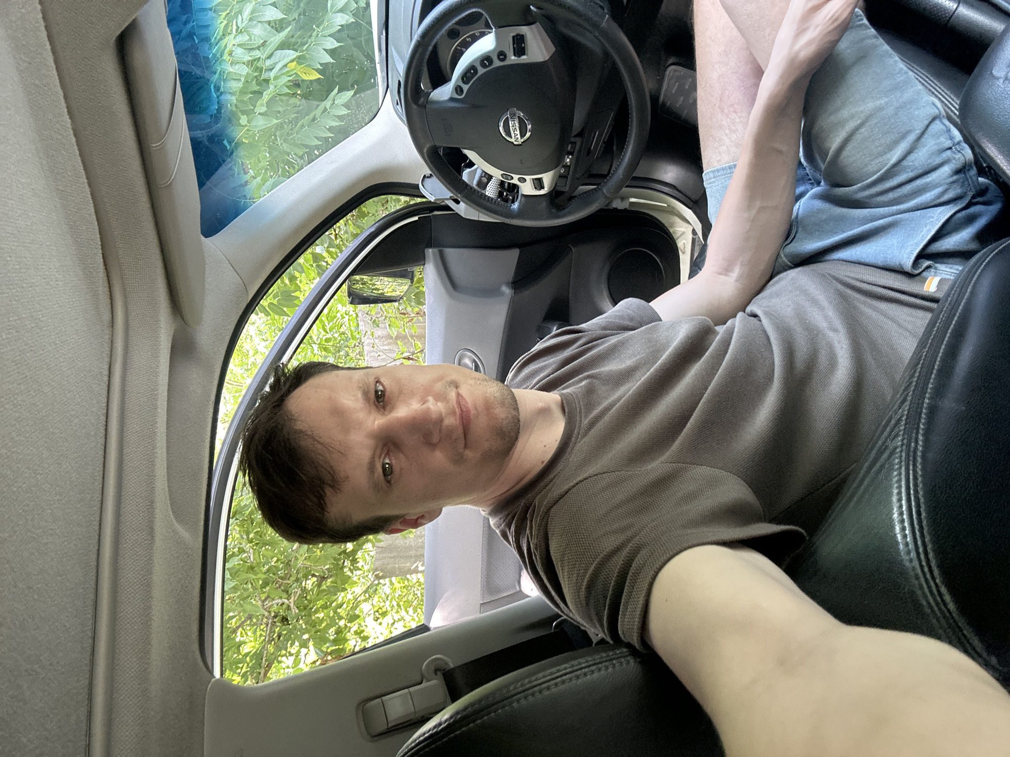 A medium shot portrait of a man in his 30s, seated in the driver's seat of a car, taken from a slightly elevated angle looking down. He has fair skin, short brown hair, and is wearing a short-sleeved brown t-shirt and light blue denim shorts. The man is looking directly at the camera with a neutral expression.
The interior of the car is visible, including the black leather driver's seat, part of the dashboard, and the steering wheel with a Nissan logo. The driver's side door is open, revealing a background of lush green trees and foliage, suggesting an outdoor setting. The lighting indicates it is daytime, with natural light illuminating the scene. The overall environment suggests the car is parked in a residential area or a place with abundant greenery. The man is believed to be located in Makiv, Ukraine.