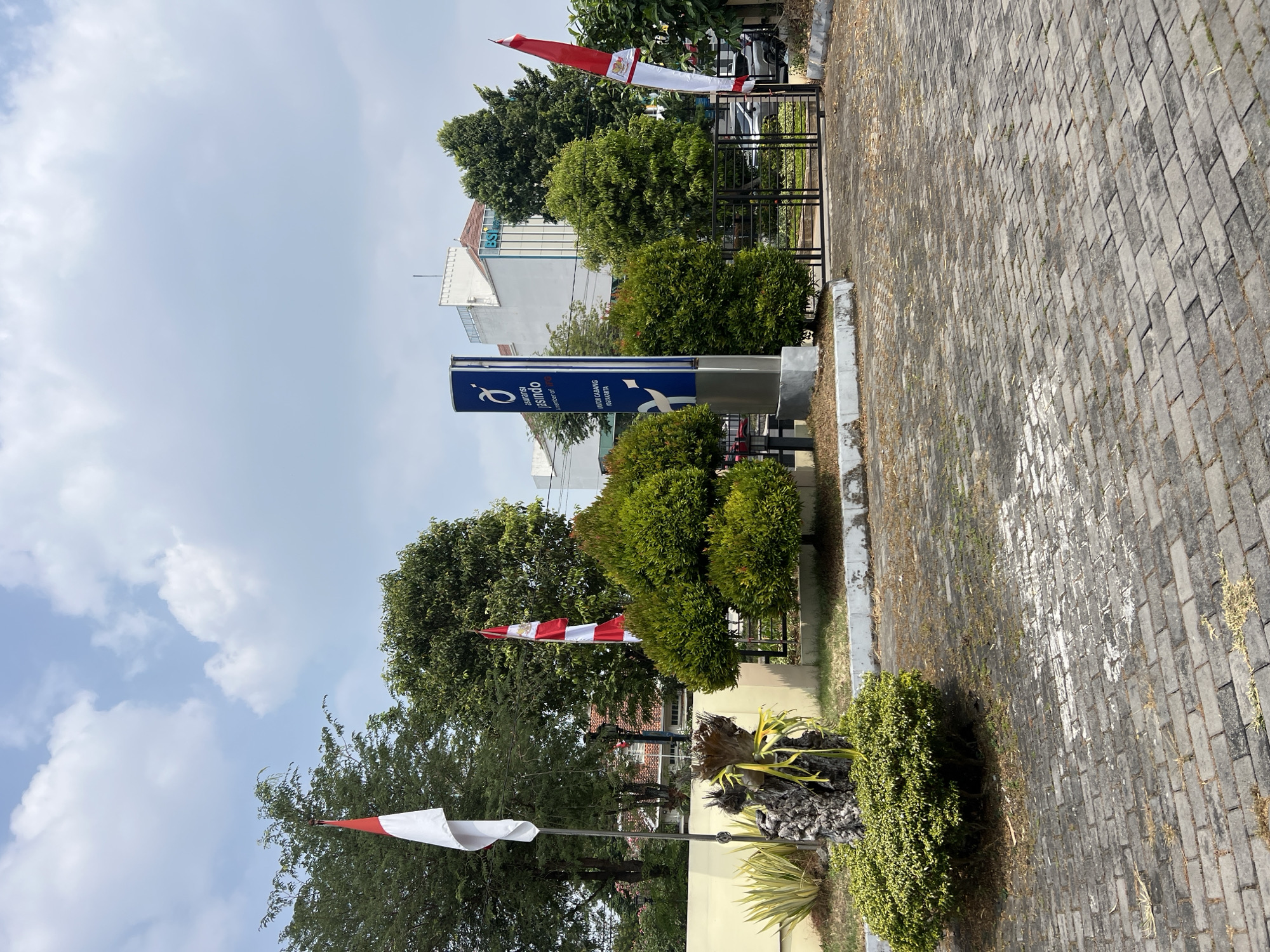The image depicts the exterior of the Jasindo Insurance branch in Yogyakarta, Indonesia. In the foreground, a paved area is visible, constructed from interlocking gray bricks. To the left, a decorative arrangement includes a large, weathered piece of wood or trunk with yellow ribbon tied around it, surrounded by greenery. Two Indonesian flags are prominently displayed: a white and red flag on a tall pole on the left, and a similar flag with a crest on a shorter pole on the right.
A blue sign with white text and a stylized logo stands near the center, reading "Asuransi Jasindo" and below that, "KANTOR CABANG YOGYAKARTA" and "A member of MIND ID". Behind the sign, a modern building with white facade and windows is visible. Several lush green trees and manicured bushes are scattered throughout the scene, creating a well-maintained outdoor space. A metal gate is positioned in the middle ground, leading to what appears to be a parking area or further grounds. The sky above is a mix of blue with scattered white clouds.