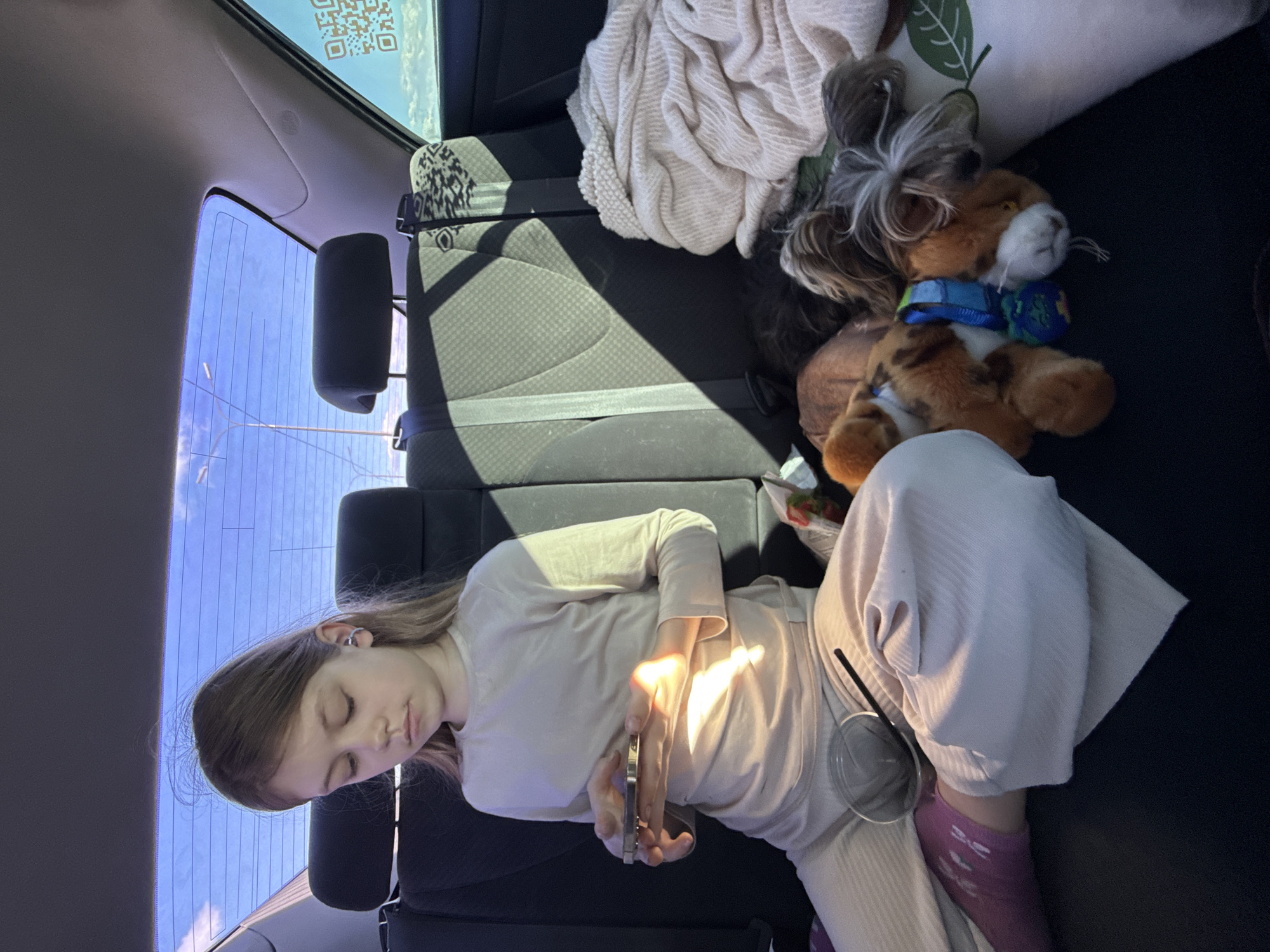 A young female is recumbent in the back seat of a vehicle. She is holding a mobile phone in her hands, her eyes are closed. She is wearing light colored clothes and lilac socks. Next to her is a clear plastic cup with a straw, a stuffed toy dog, a leaf-patterned pillow, and a light colored blanket. The vehicle is presumably moving, as indicated by the blurred view of the outside through the rear window. The QR code on the rear side of the vehicle indicates that the location is in the city of Losevo, Russia. The car seat has a black and gray patterned fabric.