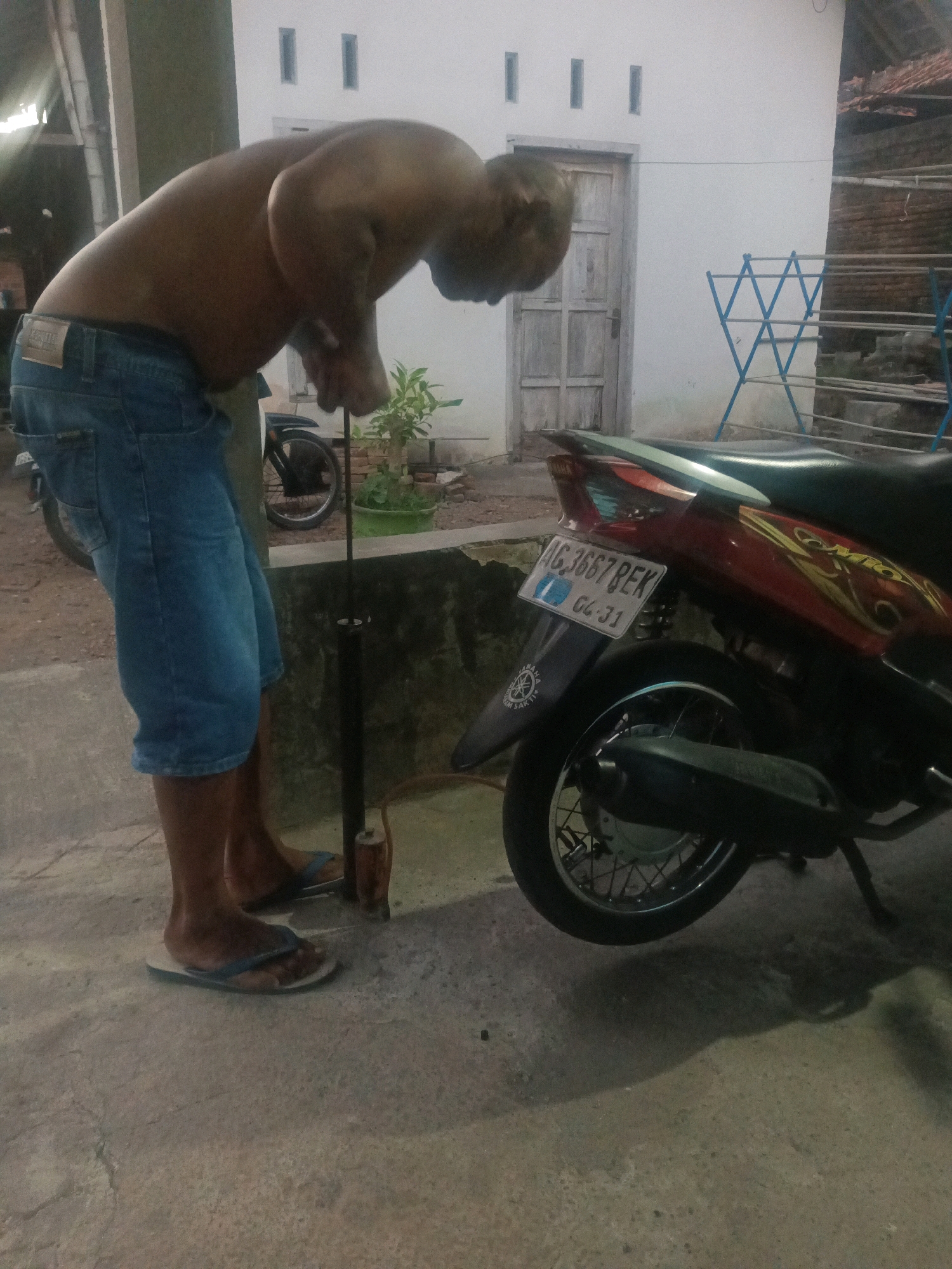 A man with a muscular build, shirtless and wearing blue denim shorts, is focused on pumping the tire of a red and black scooter. He is standing on a concrete surface, possibly a driveway or yard, in what appears to be an outdoor setting. The scooter is parked to his right, with its license plate clearly visible. Behind him, a white building with a wooden door and small rectangular windows suggests a residential area. A drying rack and some brickwork are also visible in the background, along with a small potted plant. The lighting suggests it is either late afternoon or early evening, casting shadows on the ground. The man's posture indicates he is engaged in a task that requires effort and concentration. The overall scene is domestic and casual, hinting at a routine activity.
Visible text: The license plate on the scooter reads "AG 3667 BEK" and "04-31". There is also a logo on the scooter's fender with text that appears to be "YAMAHA" and possibly a location or model name below it, though it is difficult to read clearly. A label on the man's shorts is also visible, but the text is not legible.