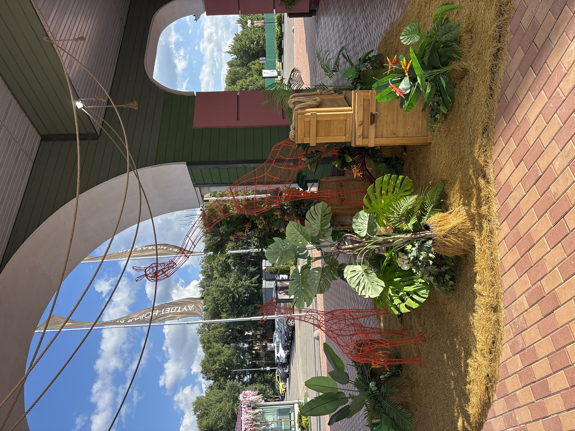 The image depicts an outdoor decorative display located at the shopping center "Pokrovskoe" in Pokrovskoe, Russia. The display features two red wireframe giraffe sculptures and artificial tropical plants, arranged on a bed of straw. Two wooden crates are stacked in the center of the display, with a coiled rope placed on top of the upper crate. The background shows an archway leading to a parking lot with several cars and trees. The sky is blue with scattered clouds. The flooring is a patterned brick walkway. Flags are visible in the background, bearing Cyrillic text. The scene appears to be set up for an event or as a permanent decorative feature.