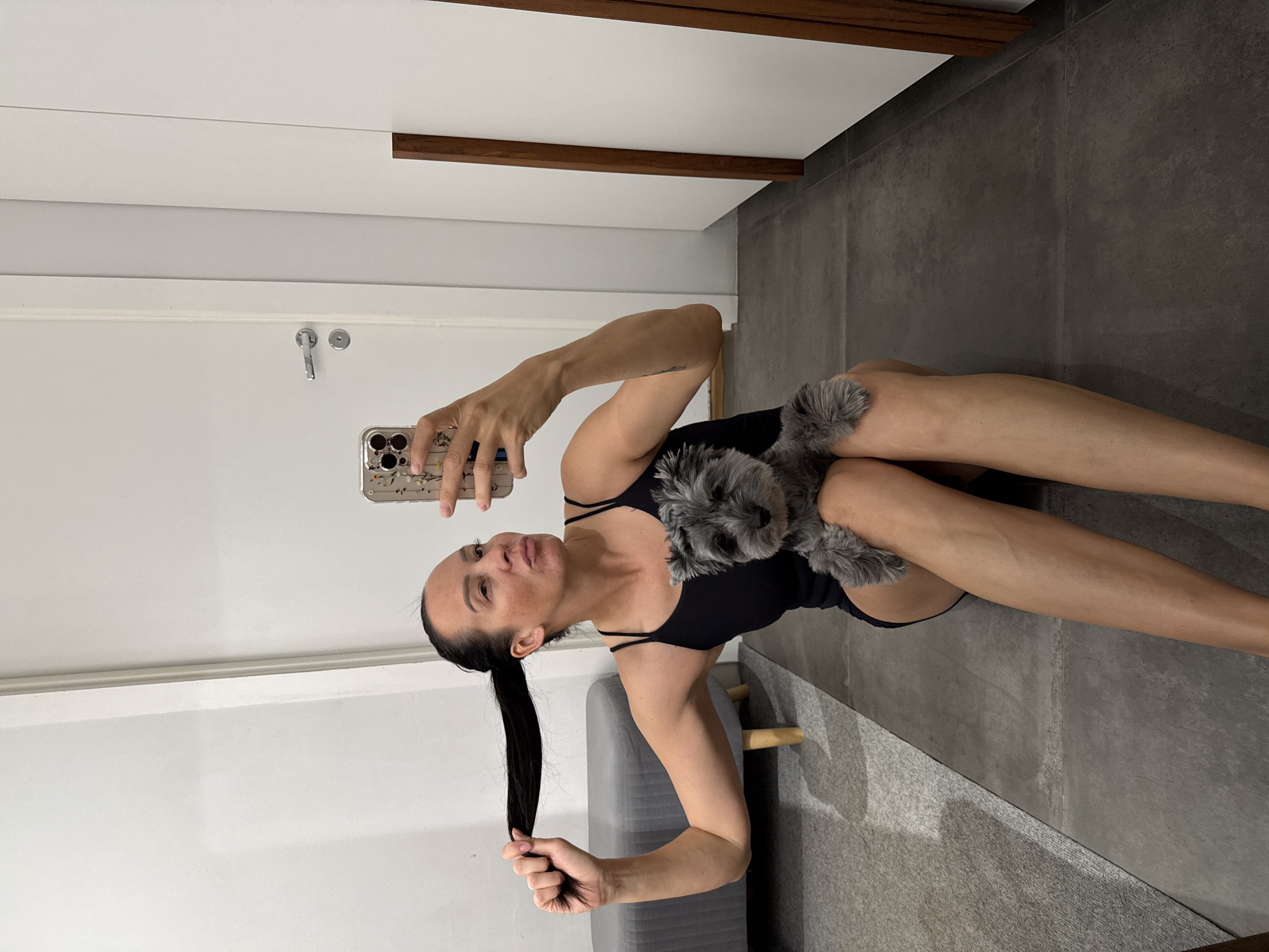 A young woman with dark hair pulled back into a ponytail is sitting on the floor, holding her hair and a small grey dog. She is looking at her phone, which she is holding up to take a selfie. The woman is wearing a black tank top. The dog is on her lap, and its paws are resting on her legs. The background is a room with white walls and a door. There is also a grey ottoman and a rug on the floor. The image appears to have been taken indoors, likely in a residential setting. The lighting is bright, suggesting the use of natural light or a flash. The woman's facial expression is playful, and the dog appears calm. The setting does not provide any specific clues about the location within the city of Chaikovsky, Russia, as it depicts a generic indoor environment.