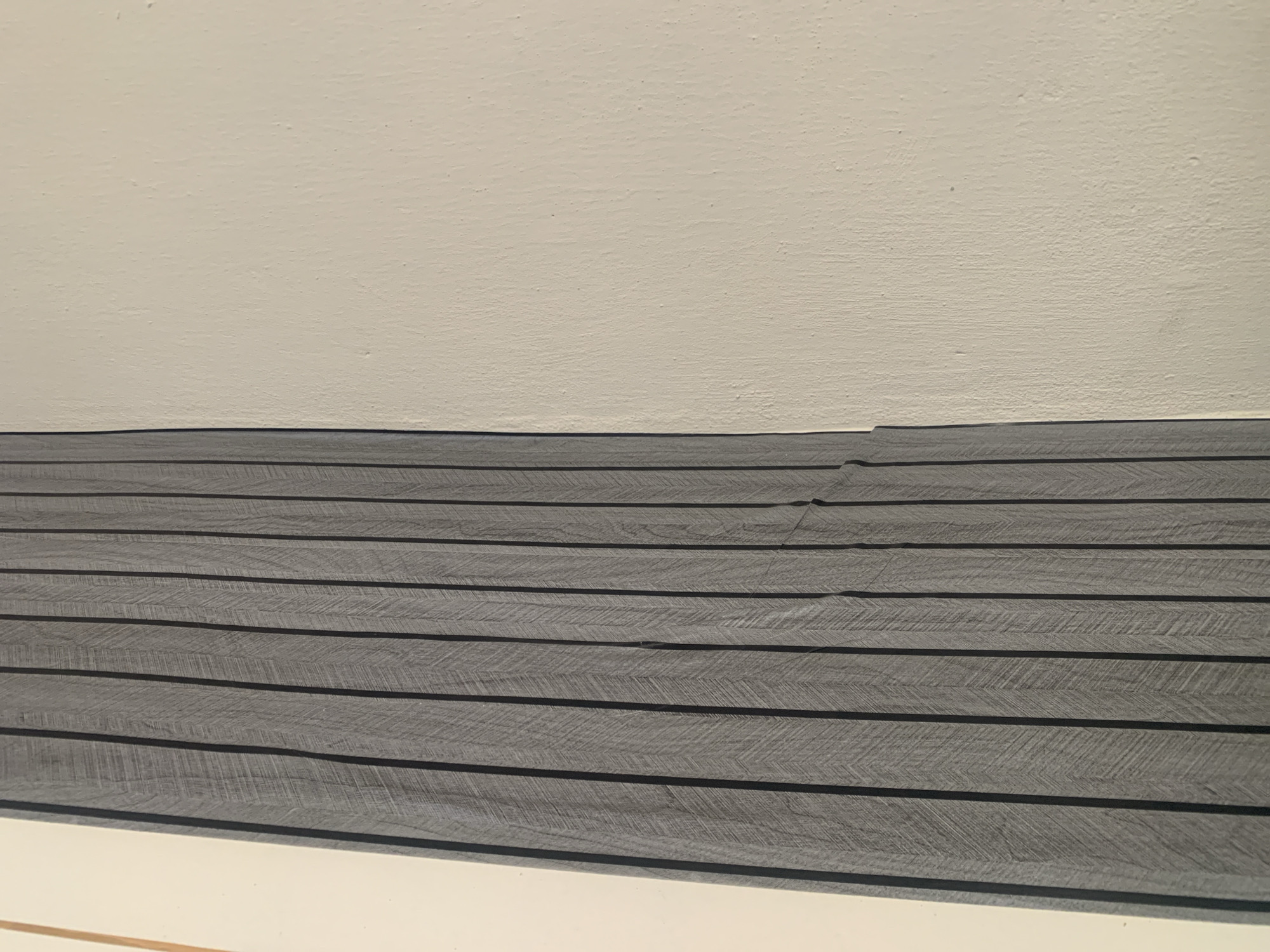 A close-up, head-on shot shows a portion of a wall with vertical wooden slats on the left and a painted wall on the right. The wooden slats are gray with a subtle wood grain texture and are arranged in parallel, creating a ribbed effect. The painted wall is a light, off-white color and appears to have a slightly textured surface. The image is lit from the right, casting shadows that emphasize the texture of the slats and the wall. No people, objects, or actions are visible. The scene is a close-up detail of interior architectural elements.