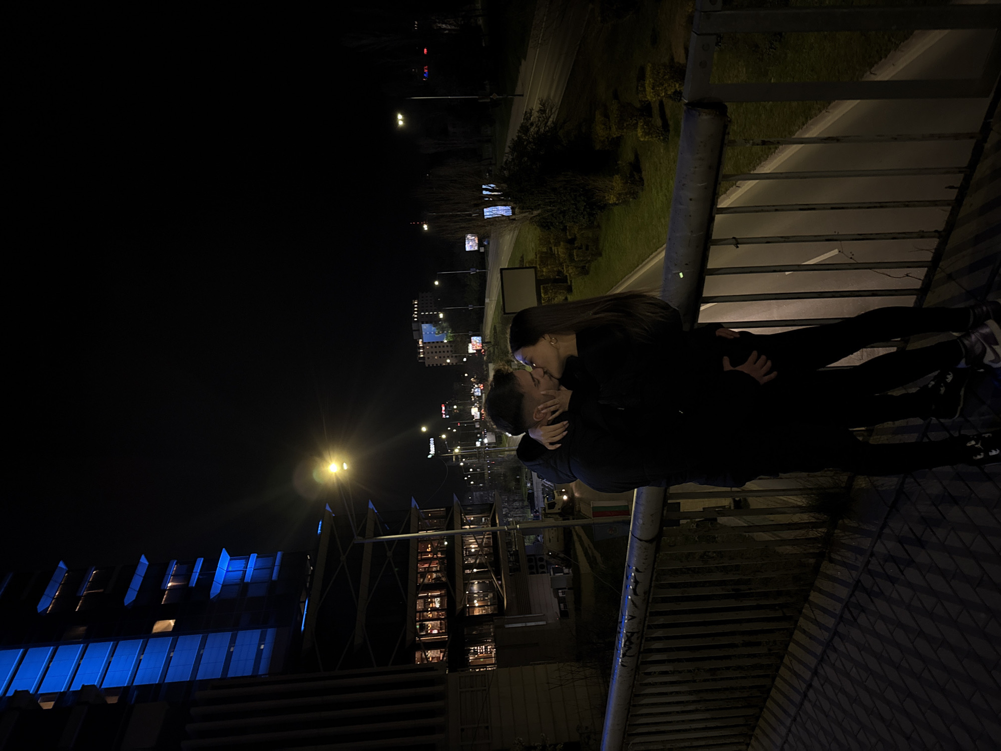 A low-angle, medium shot at night depicts two people embracing and kissing. The location appears to be a pedestrian bridge or elevated walkway in Sofia, Bulgaria. The people are standing against a metal railing. The background includes city lights, illuminated buildings, and trees. A large building with blue lighting is visible in the lower left. A road with streetlights is visible behind the walkway.