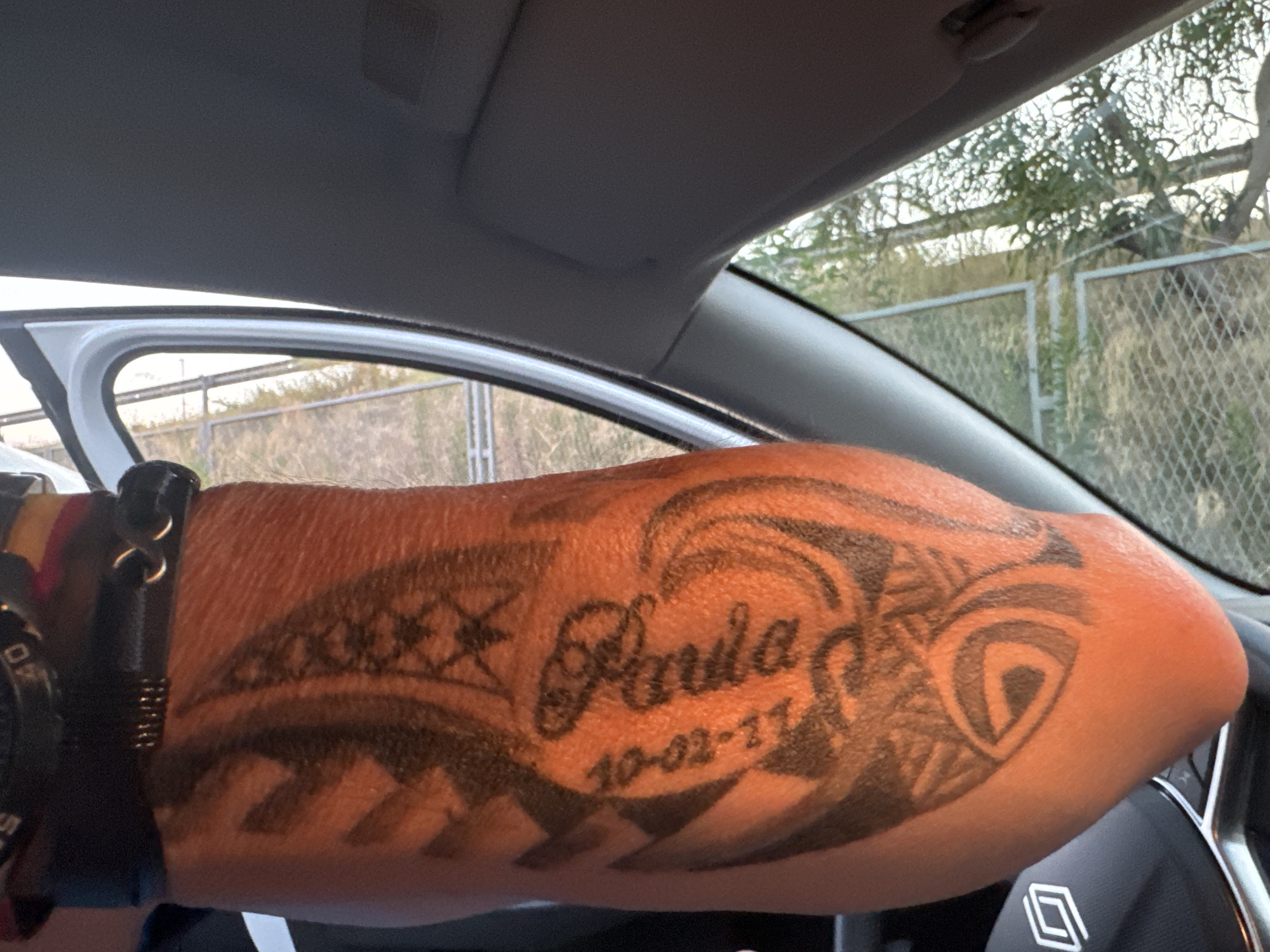 The image shows a person's forearm with a tattoo. The tattoo appears to be tribal in design, with a name "Paula" and the date "10-02-11" inked below it. The person is inside a vehicle, as indicated by the car's interior visible in the background, including a steering wheel and part of the dashboard. The exterior view shows a fence and trees, suggesting an outdoor environment. The lighting suggests it is daytime.