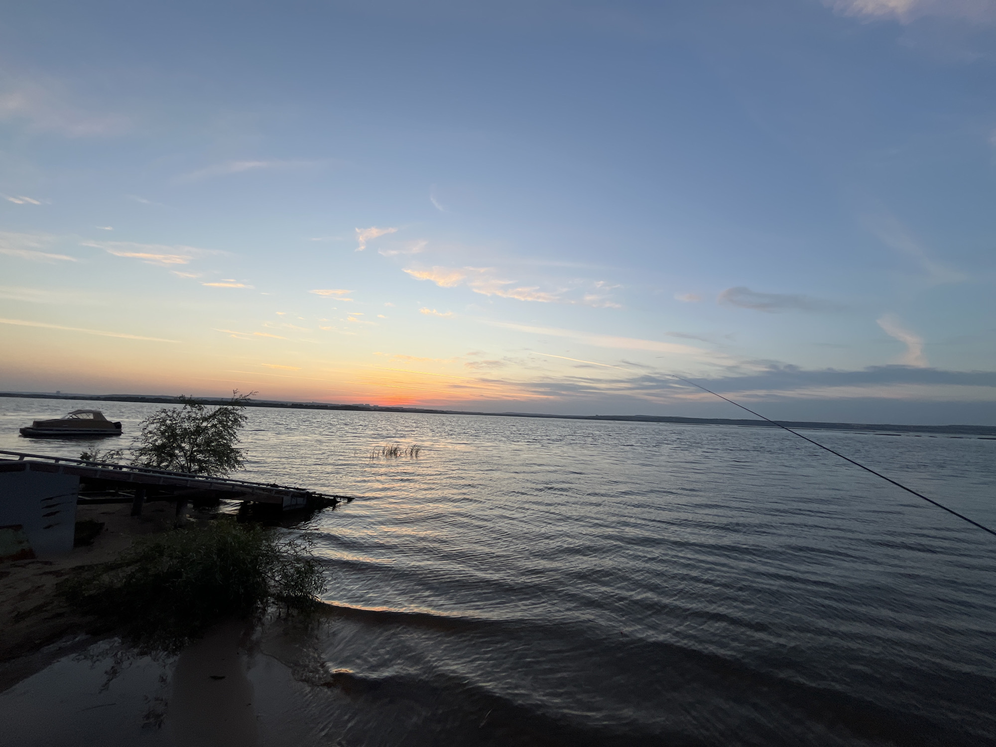 The image depicts a serene sunset over a large body of water, likely a river or lake, near the settlement of Shumeika, Russia. The sky transitions from a soft orange and pink hue at the horizon, where the sun has just set, to a clear blue overhead, with scattered wispy clouds. The water is calm, with gentle ripples reflecting the fading light.
In the foreground, a sandy shoreline is visible, with some vegetation and a small, somewhat dilapidated wooden pier. A small boat is moored to the left of the pier, partially submerged. A single fishing rod extends from the right side of the frame, its tip barely visible against the sky, suggesting an individual is fishing. The overall atmosphere is peaceful and tranquil, characteristic of a rural evening.