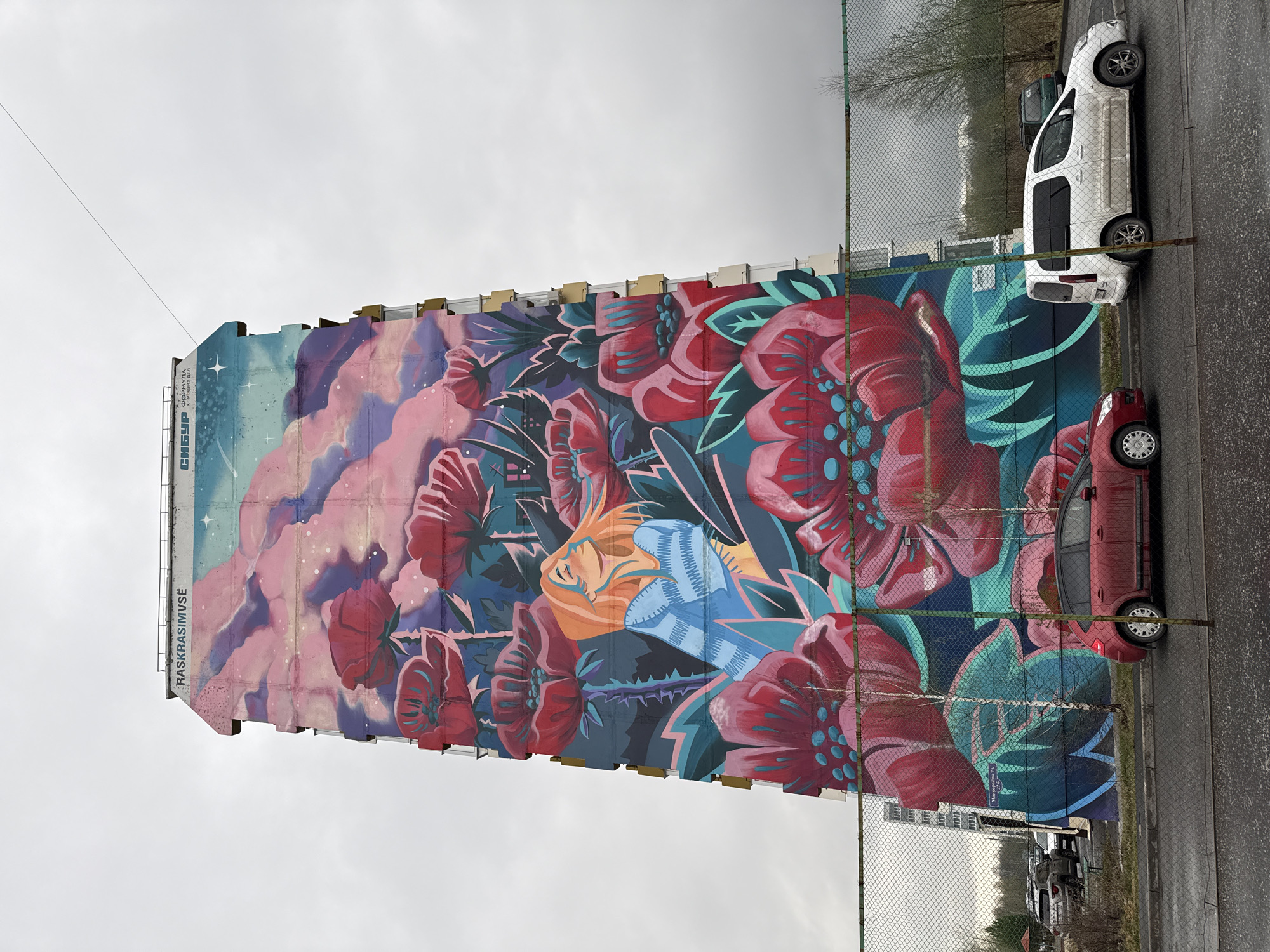 A mural adorns the side of a building in Tobolsk, Russia. The mural depicts a dreamlike scene with vibrant flowers, and a face. The name of the town can be found on a street sign. The sky is overcast. A white car and a red car are parked along the side of the building. A fence runs along the edge of the mural and adjacent parking area.