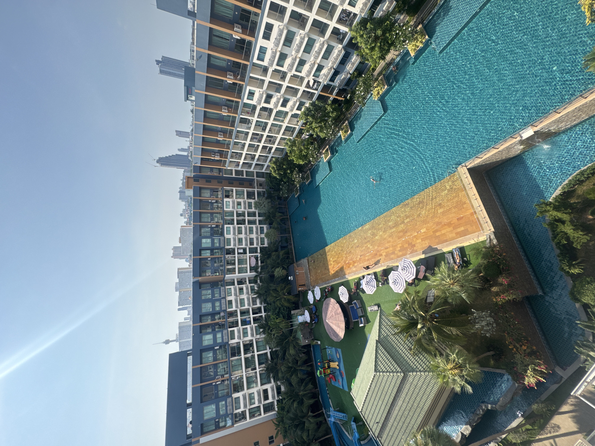 The image shows an elevated view of a residential complex in Pattaya City, Thailand. The complex consists of several high-rise buildings with a grid-like facade of balconies and windows. A large swimming pool complex spans the length between the buildings, featuring tiled pools of varying depths and layouts. Trees and landscaped gardens line the pool areas. Sun loungers and striped umbrellas are arranged on a grassy patch. A children's play area with slides and climbing structures is visible. The sky is light blue with a lens flare. City buildings are visible in the distance.
