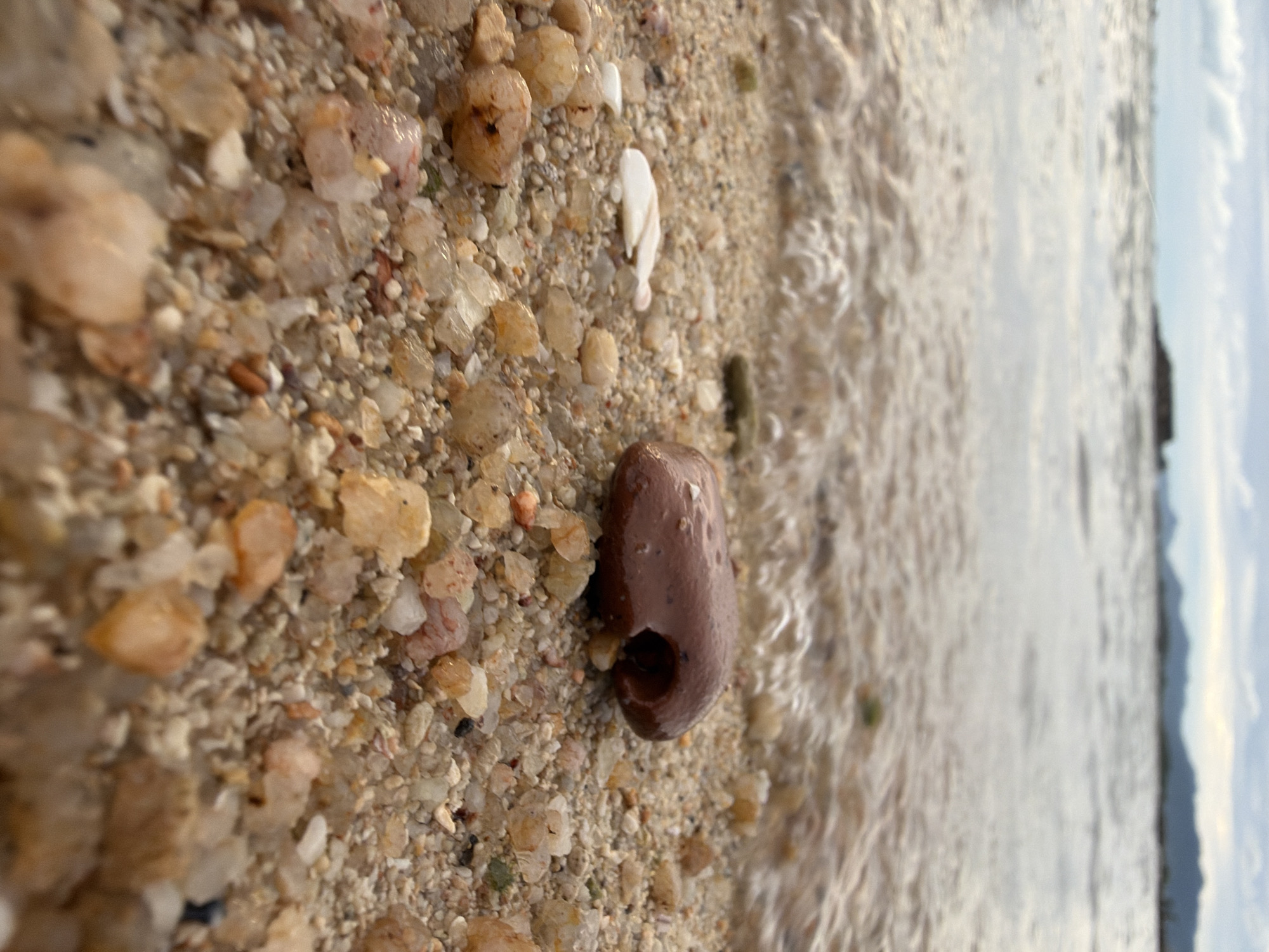 The image is a close-up, low-angle shot of a beach in Ko Samui District, Thailand. The foreground is dominated by wet sand and various pebbles, ranging in color from light brown to translucent amber and white. A single, smooth, dark brown stone with a hole in its center is prominently placed on the sand. A small, white shell fragment is visible to the left of the stone. The middle ground shows the edge of the water where small waves are gently washing ashore, creating foamy ripples on the sand. In the background, the sea stretches out towards a hazy horizon with distant islands or landmasses visible under a cloudy sky. The overall scene depicts a natural, serene beach environment.