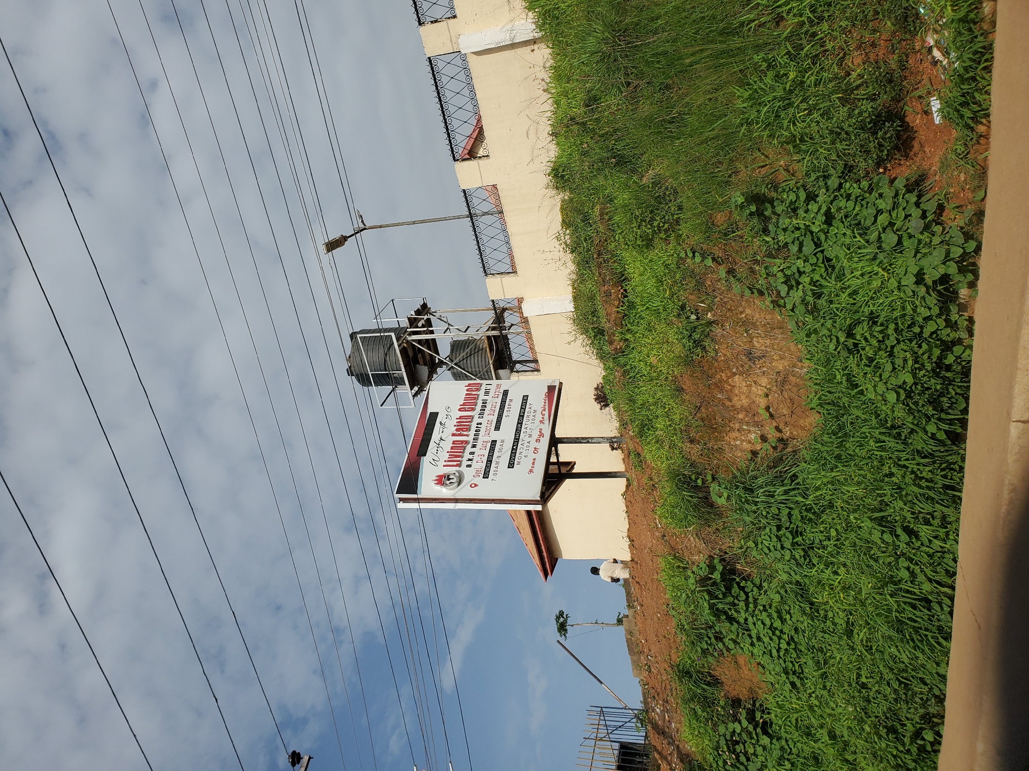 In Bukuru, Nigeria, an outdoor scene depicts a billboard advertising "Living Faith Church," also known as "a.k.a winners chapel int'l." The billboard, situated in front of a building and a hillside covered in lush green vegetation, provides service times for Sunday services (7:00 AM - 9:00 AM and prayer meeting 5:00 PM) and weekday prayer (Monday to Saturday, 6:30 AM - 7:30 AM). The church's motto, "Home Of Signs And Wonders," is also visible. Adjacent to the building, a water tank stands on a metal structure. A person wearing a white garment walks away from the billboard, heading up the hill. Overhead, power lines stretch across a cloudy sky. The environment appears to be a roadside with a sloping terrain.