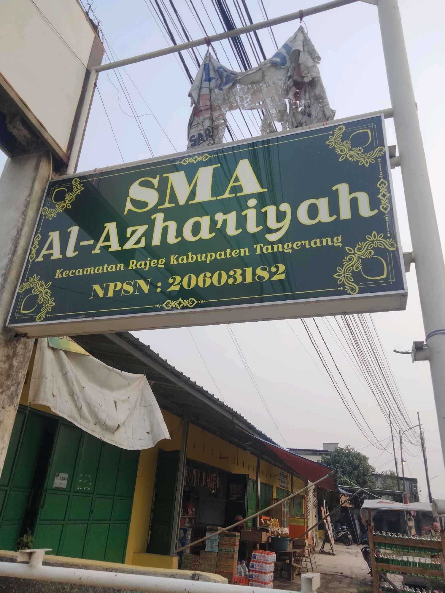 The image shows a sign for "SMA Al-Azariyah" in the city of Suka Sari, Indonesia. The sign is green with yellow lettering and decorative elements. Below the school name, it states "Kecamatan Rajeg Kabupaten Tangerang" and "NPSN: 20603182". The sign is attached to a pole and is partially obscured by wires and a tattered piece of fabric hanging above it. In the background, there is a building with green metal doors and a yellow facade, with a white tarp stretched over part of it. There are also some stacks of bottled water and other goods visible near the building. The sky is overcast.