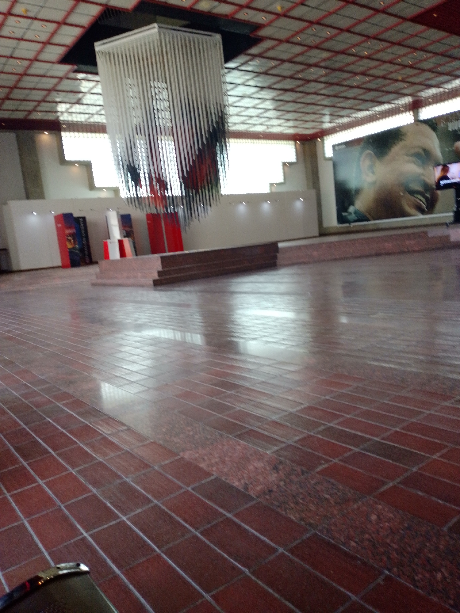 The image shows the interior of a large hall, likely a public space, located in Puerto La Cruz, Venezuela. The hall features a tiled floor in shades of red and brown, a geometric grid-patterned ceiling, and an elaborate chandelier-like structure hanging from the ceiling. There is a low stage area with steps. On the wall to the right is a large portrait of a man, possibly a local figure, and a television screen. There are various display panels and structures arranged in the background. The hall appears to be empty.