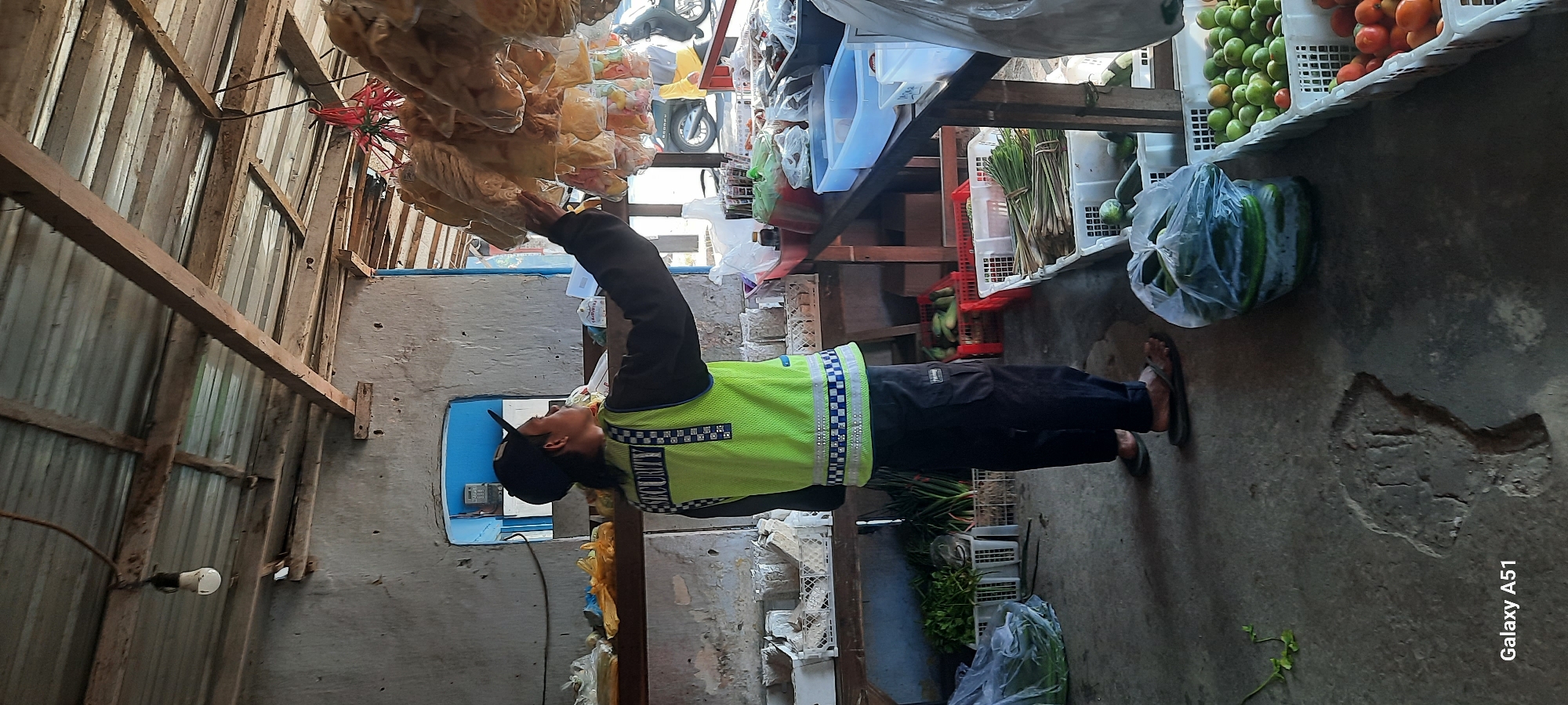 A person wearing a high-visibility vest and a baseball cap stands inside an open-air market stall in Selopuro, Indonesia. The stall has a corrugated metal roof supported by wooden beams and a rough-hewn wall. The person is reaching up to a display of packaged snacks hanging from the ceiling. In the foreground, crates of fresh produce, including green vegetables and tomatoes, are visible on the concrete floor. The scene suggests a typical rural market environment.