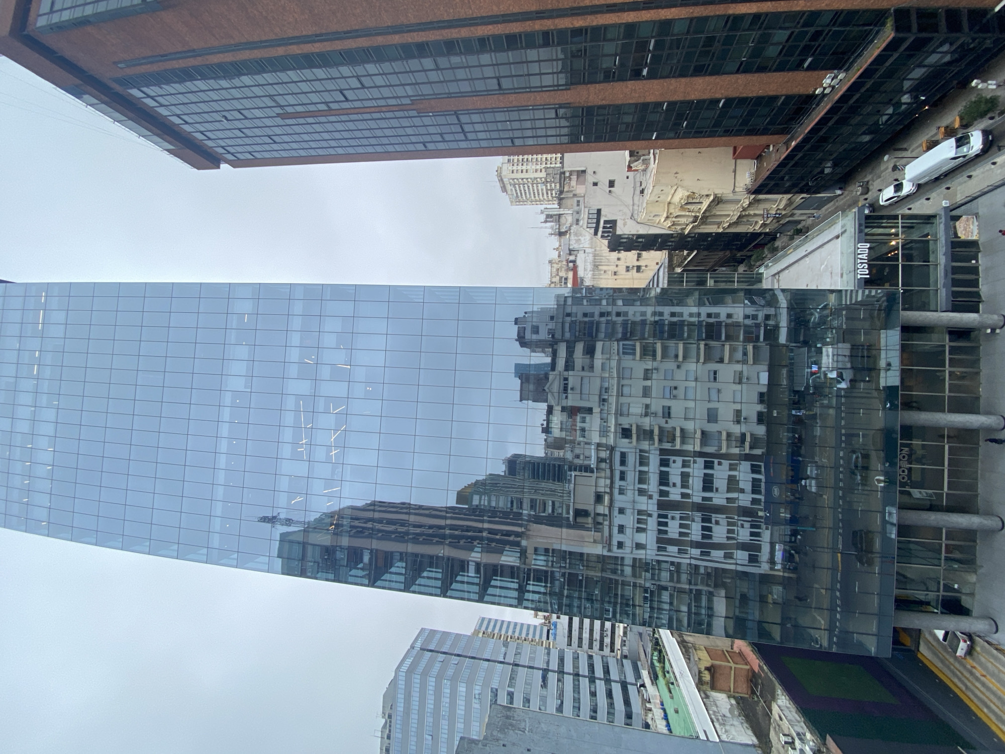 The image shows a view from an elevated position in Buenos Aires, Argentina, looking towards a glass-faced skyscraper. The glass reflects the surrounding buildings and a cloudy sky. To the right, a street scene is visible, including a Tostado cafe. Several buildings of varying heights are present, with a brick building at the top and other contemporary office buildings on the left. Cars and pedestrians are faintly visible on the street. The general atmosphere appears to be urban and overcast.