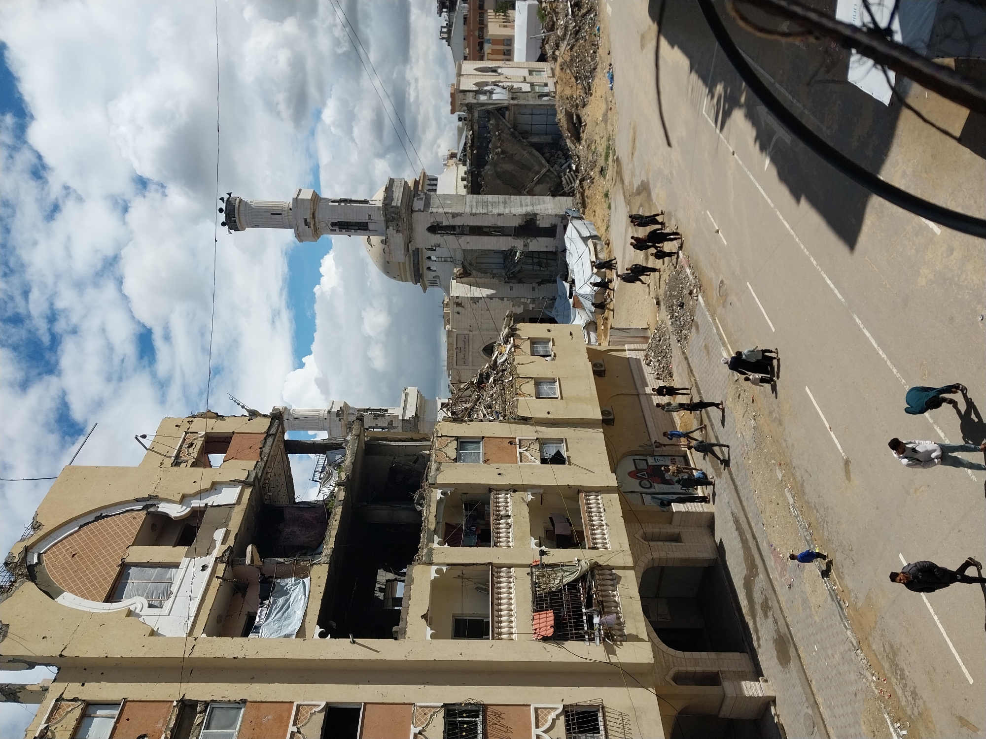 This image depicts a street scene in Khan Yunis, Palestinian Territory, showing significant destruction to urban structures under a partly cloudy sky.
On the left, a multi-story building with a light beige facade exhibits extensive damage. Multiple floors have large sections missing from their exterior walls and ceilings, exposing internal rooms and debris. The building's facade shows numerous impact marks consistent with bullet holes. Architectural features like pointed archways above windows are visible, some damaged. Balconies with white balustrades are partially intact or collapsed. White and blue tarps hang from several openings.
On the right, a prominent mosque stands heavily damaged. Its white minaret, cylindrical with a decorative top section and loudspeakers, remains mostly upright. The main mosque building beneath the dome has large sections of its walls collapsed, revealing rubble and internal structural elements. A white dome sits atop the mosque. Adjacent to the mosque, significant piles of rubble and debris are scattered across the ground, with temporary white tarpaulin shelters visible near its base.
A paved road runs through the lower half of the image, featuring white double solid and broken lane markings. A sidewalk runs along the base of the damaged building on the left, strewn with dirt and debris.
Numerous individuals are present in the scene. In the immediate foreground, three men walk on the road; one in a dark jacket moves left, while two others in lighter clothing walk right, one bending down. Further along the road and sidewalk, multiple groups of people, including adults and children, walk in various directions amidst the debris and damaged infrastructure. A child in a blue shirt walks alone near the sidewalk. Another group includes a woman in a dark abaya and hijab with a child. A large group of people gathers near the entrance of the damaged building on the left. Individuals are also seen walking in the dirt area near the damaged mosque. The overall environment indicates an active, albeit heavily damaged, urban area.